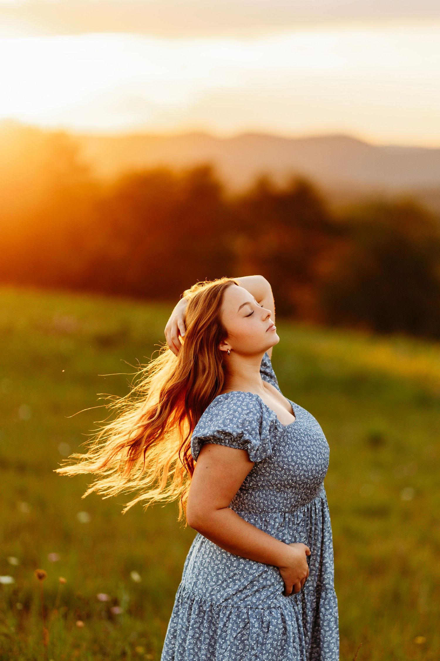 Plus size woman in blue floral dress with flowing hair at golden sunset in meadow.