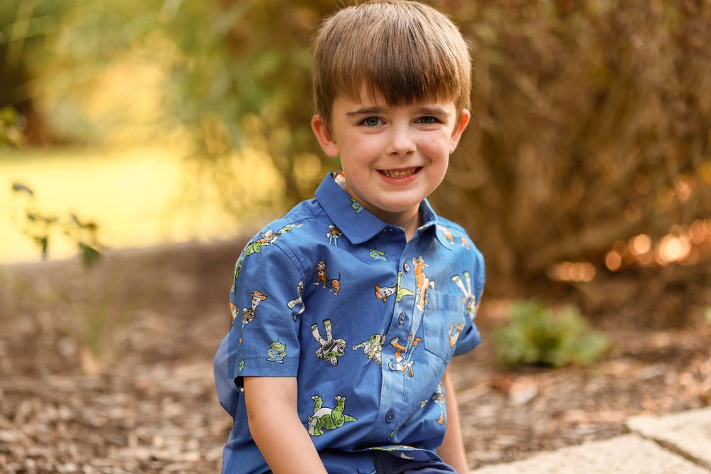 Young boy in a blue dinosaur print shirt smiling outdoors in autumn lighting.