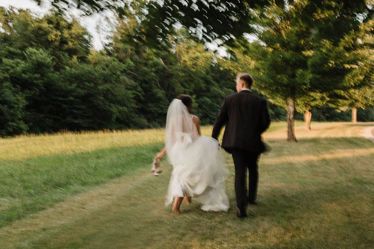 A bride and groom walking away together on a grassy path at sunset.