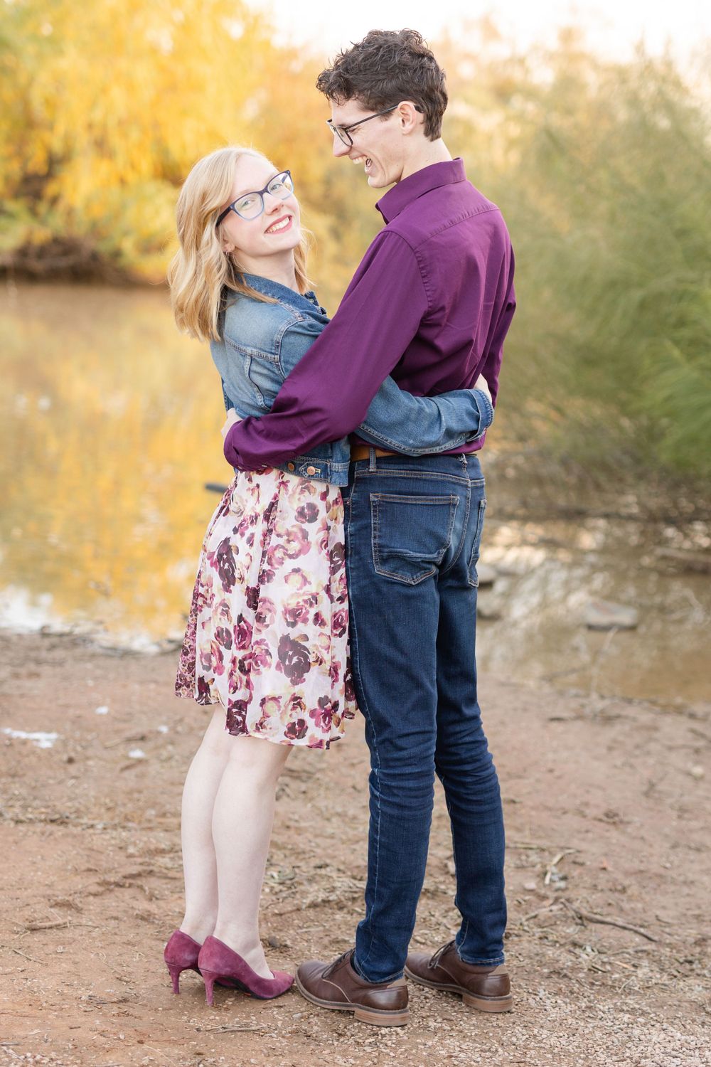 Couple in coordinated outfits embraces by waterfront during golden hour.