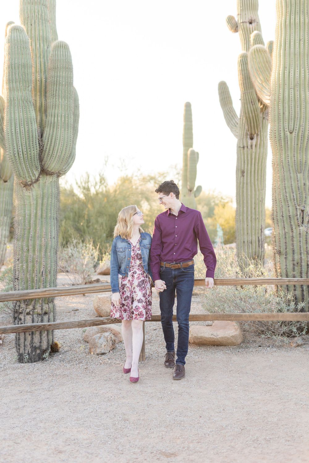 Couple walking hand in hand through desert landscape with towering saguaro cacti.