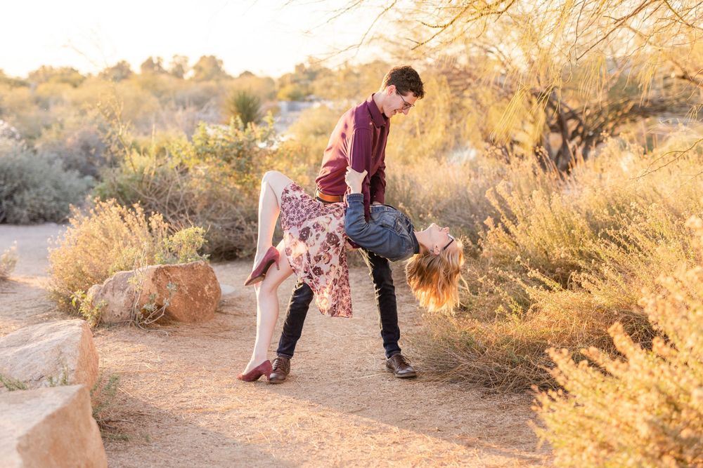 Romantic couple dips into dance pose on desert trail at sunset with shrubs and rocks in background.