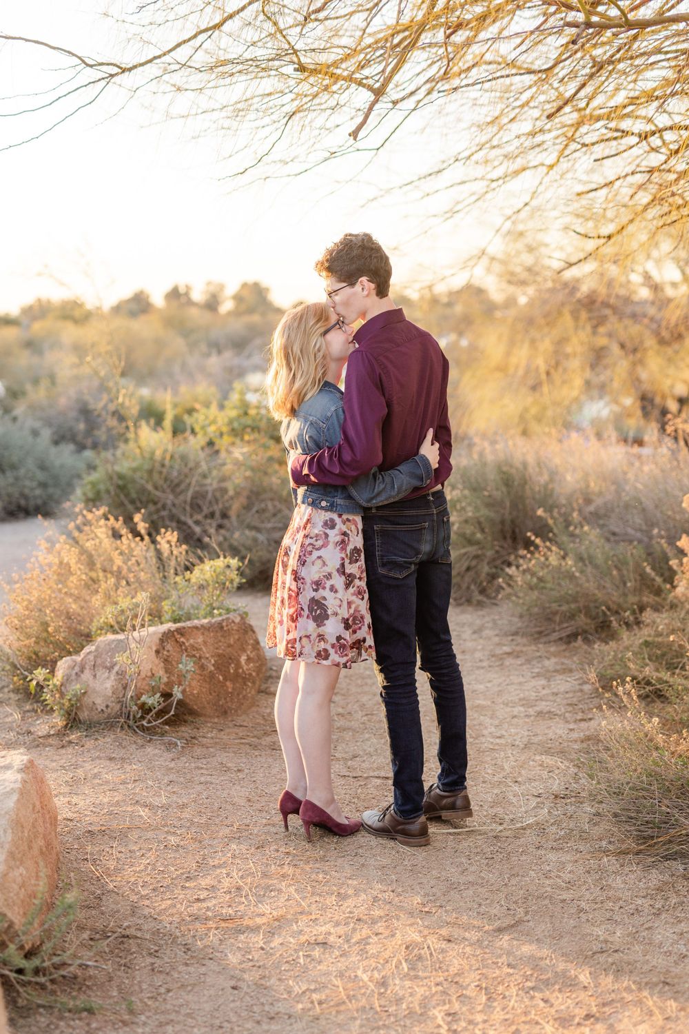 Couple shares intimate moment on desert trail during golden hour with branches overhead.