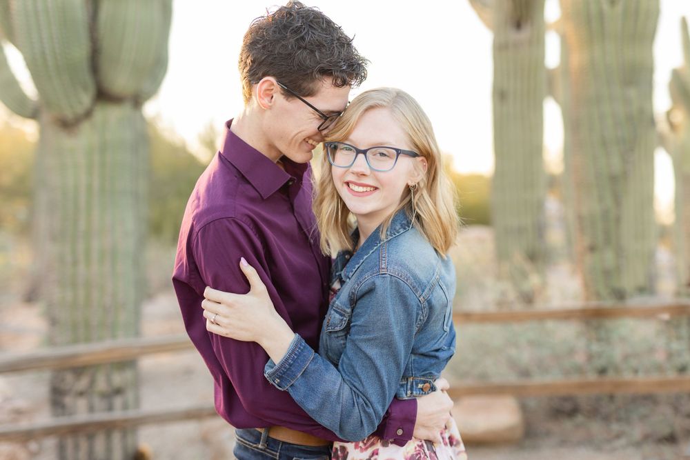 Couple in a burgundy shirt and denim jacket share tender moment among desert cacti.