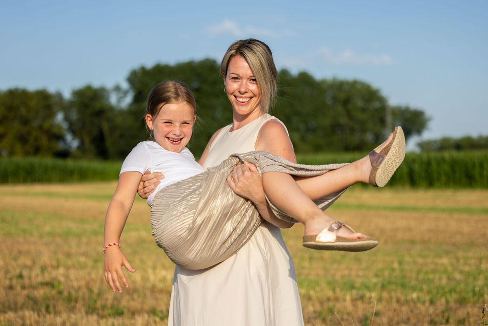 Joyful outdoor portrait of mother carrying child in cream-colored summer dress.