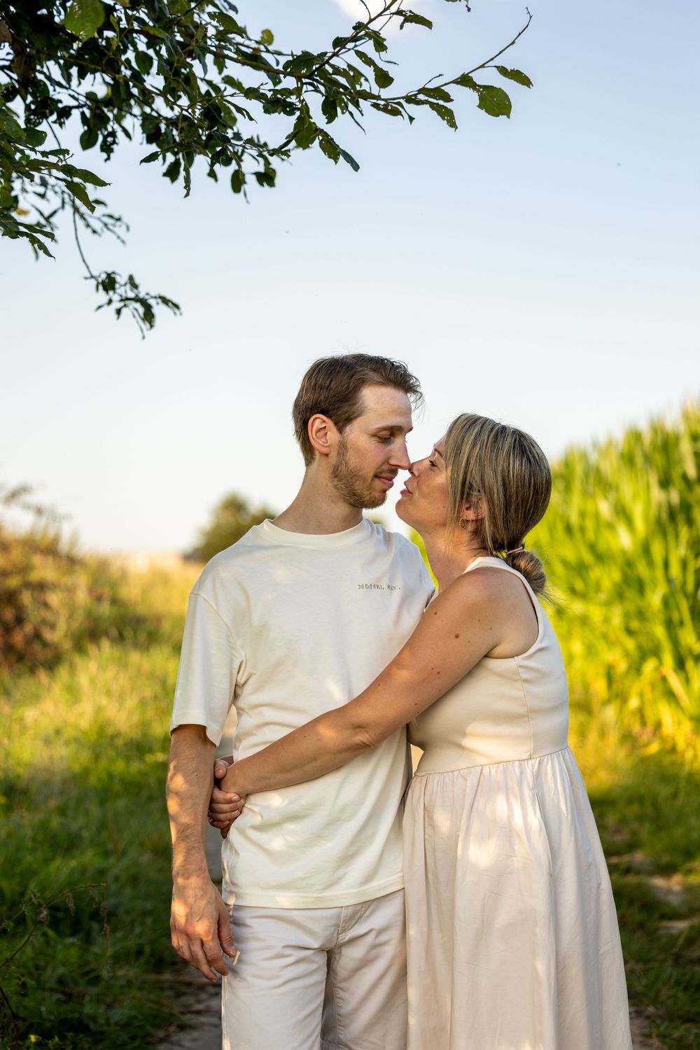 Couple in white outfits sharing an intimate moment in front of a cornfield at sunset.