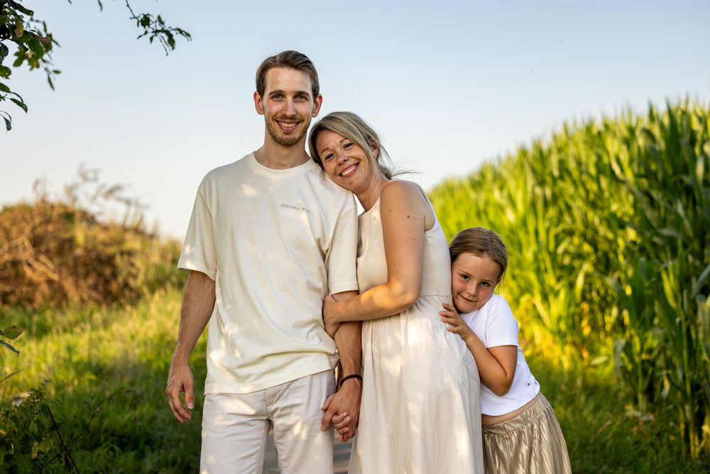 Family of three in white outfits standing together by a cornfield.