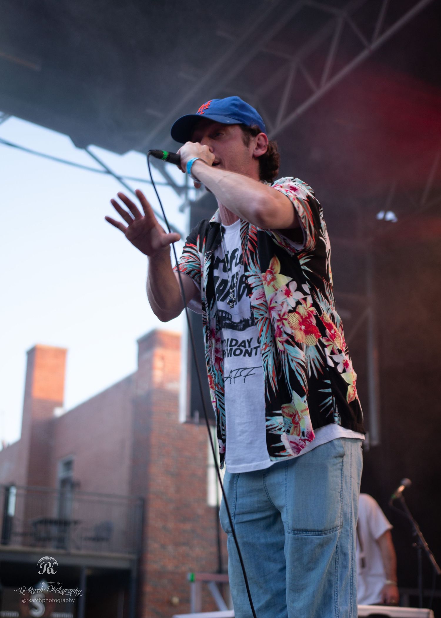Performer in floral shirt and baseball cap gestures energetically on outdoor concert stage.