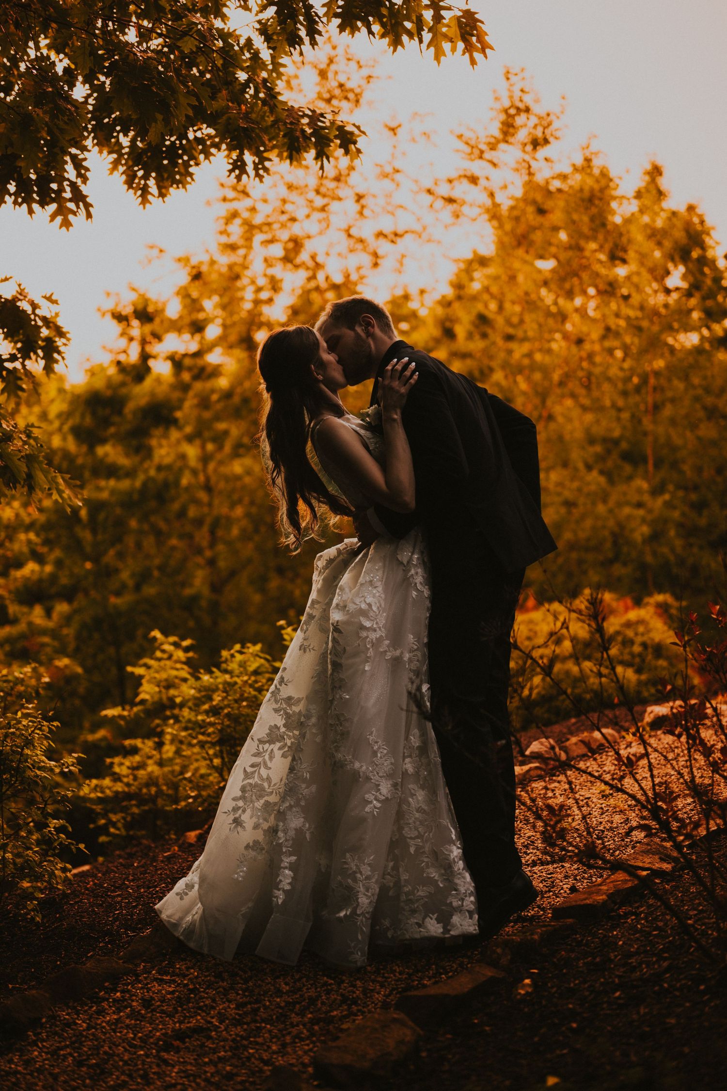 Bride and groom sharing a passionate kiss surrounded by golden autumn foliage at sunset.