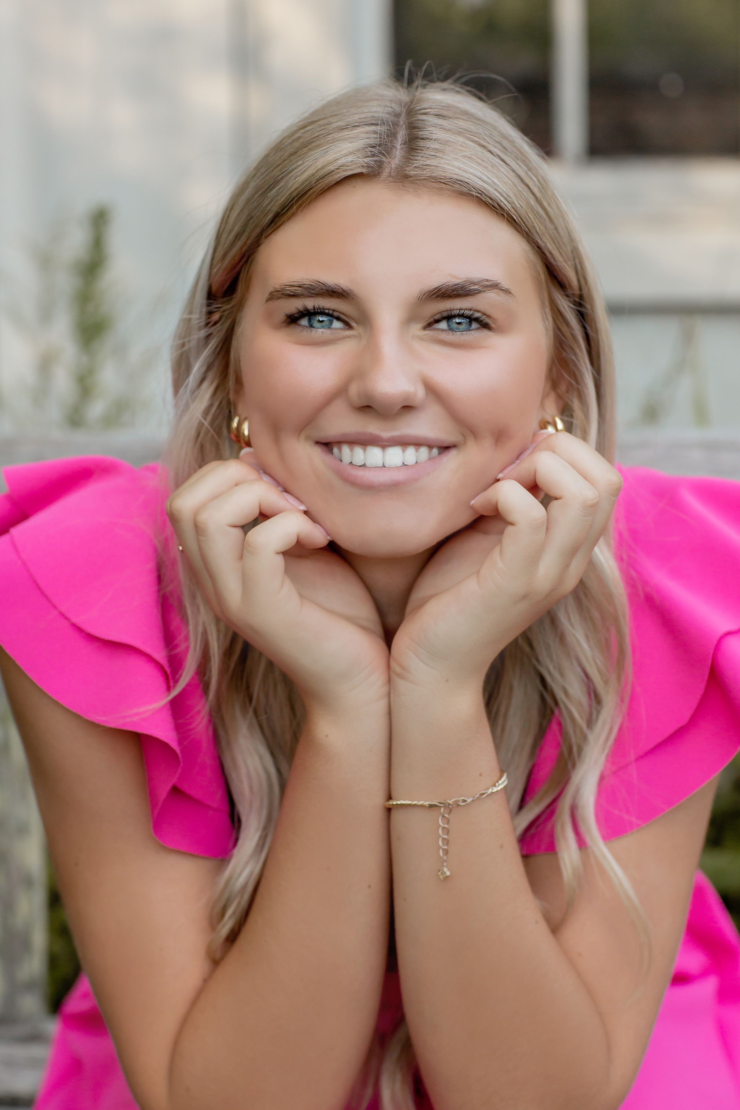 Smiling blonde woman in pink ruffled top with hands framing her face outdoors.