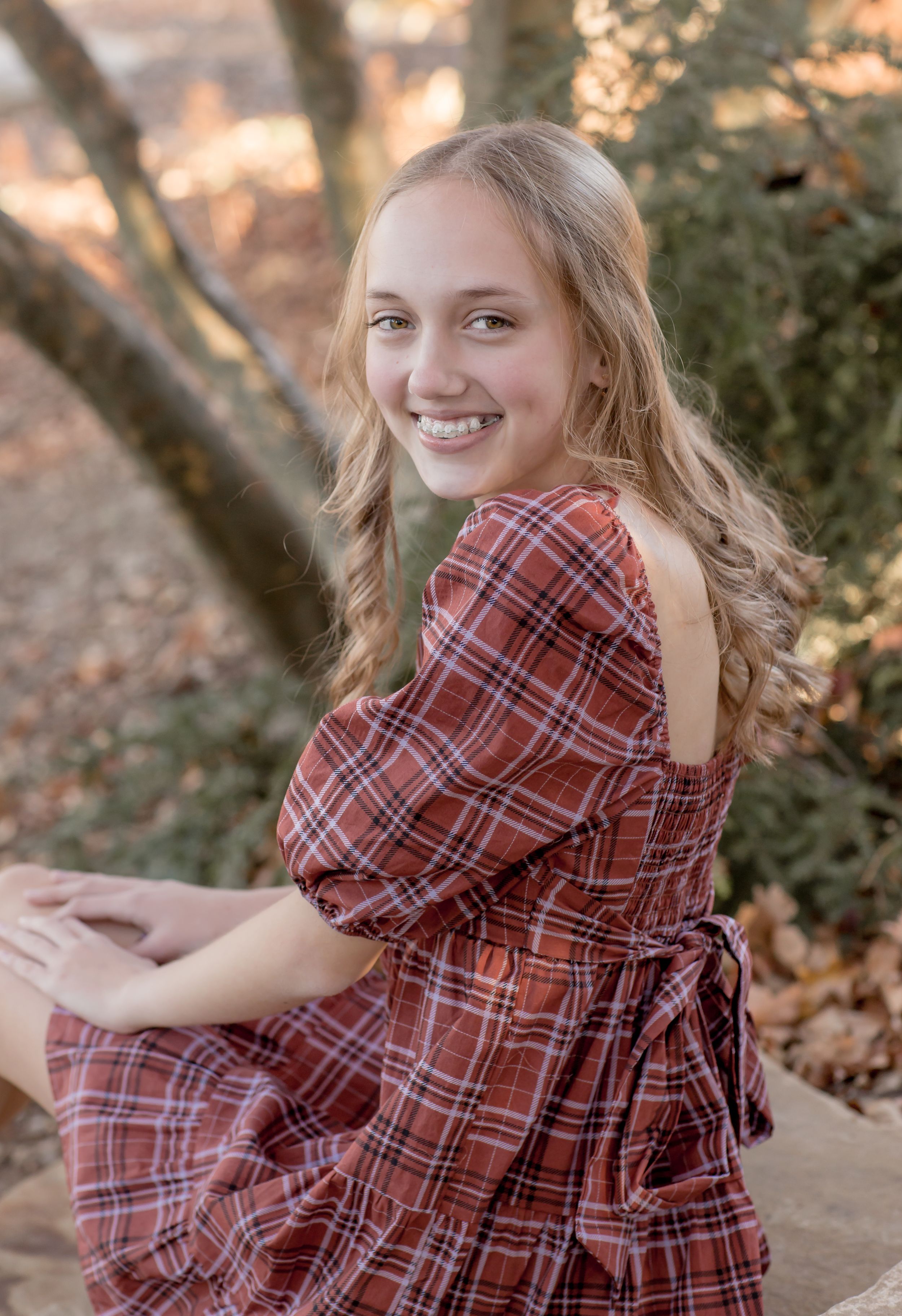 Smiling teenage girl in red plaid cold-shoulder dress posing outdoors among autumn trees and foliage.