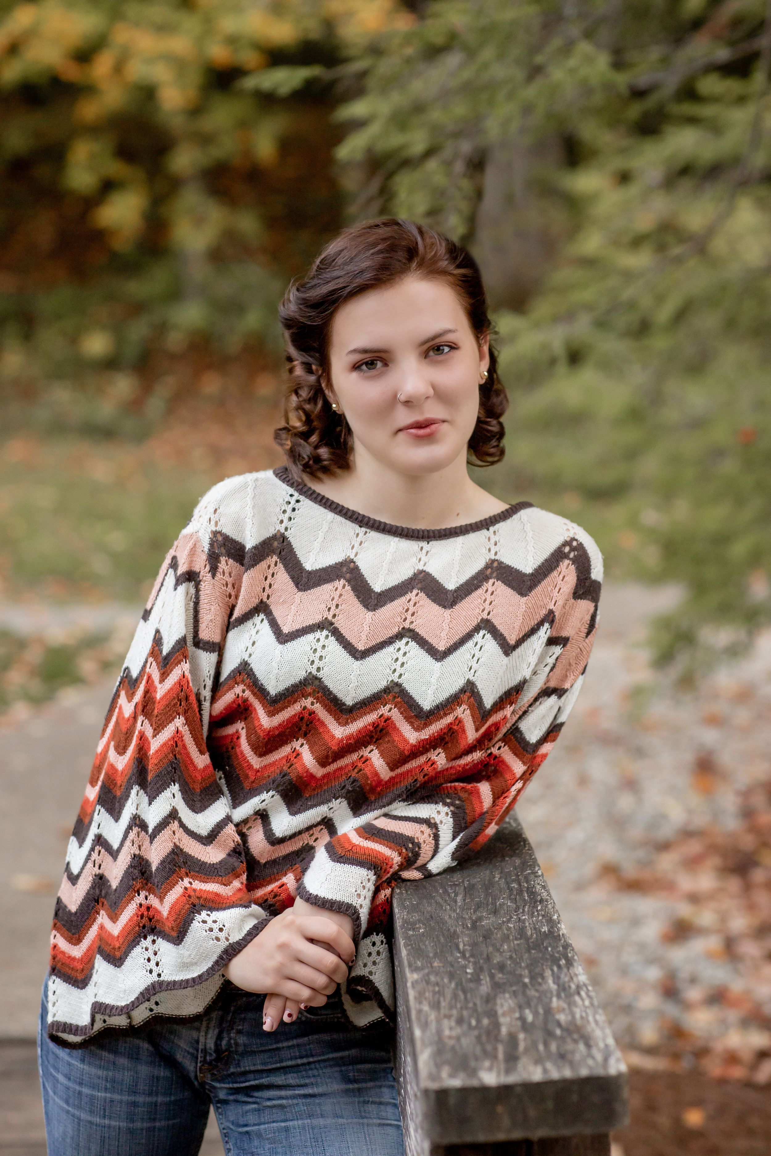 Teen girl wearing colorful chevron zigzag sweater and jeans posing outdoors in autumn forest setting.