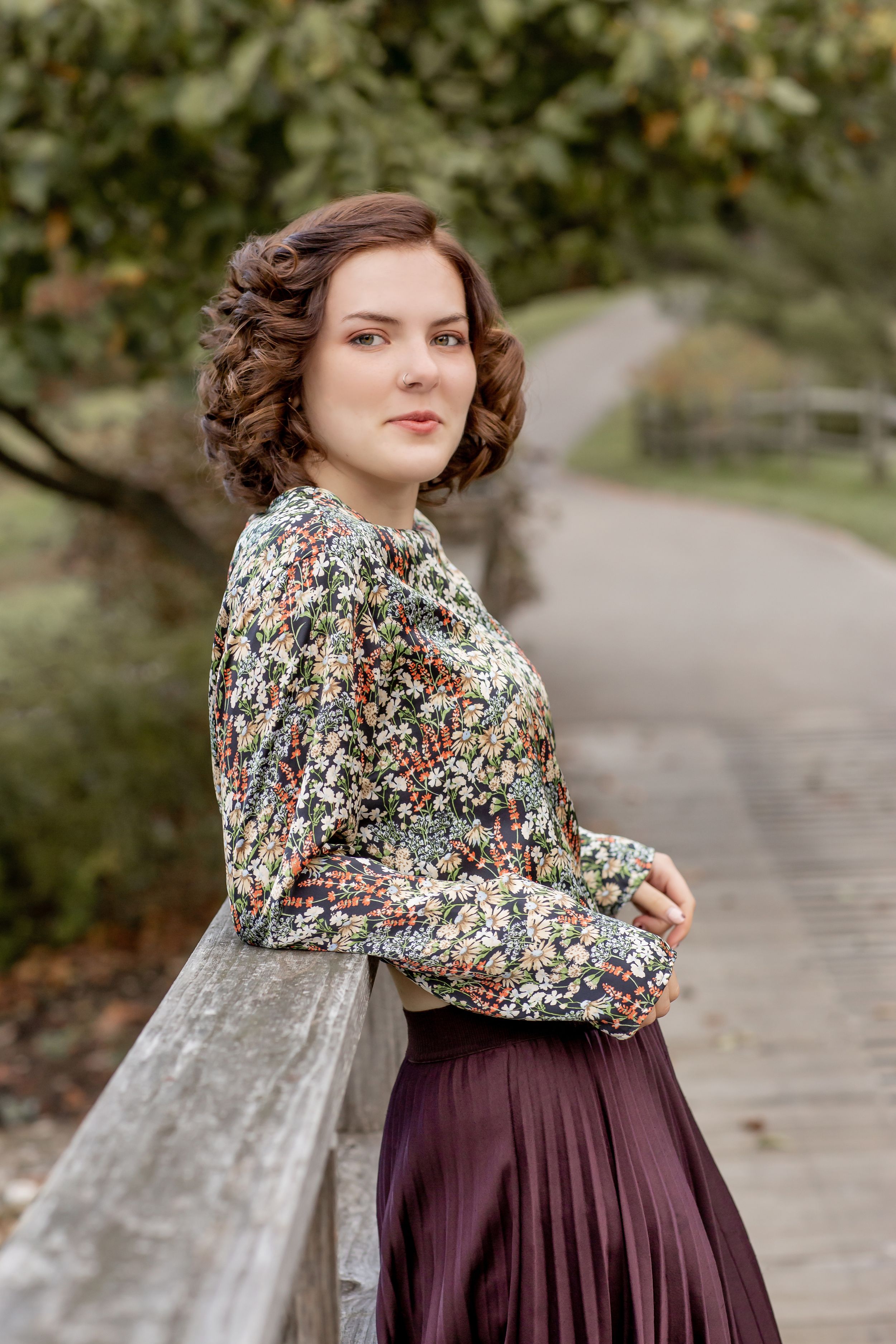 Young woman in vintage floral blouse and burgundy skirt leaning on wooden bridge railing in park.