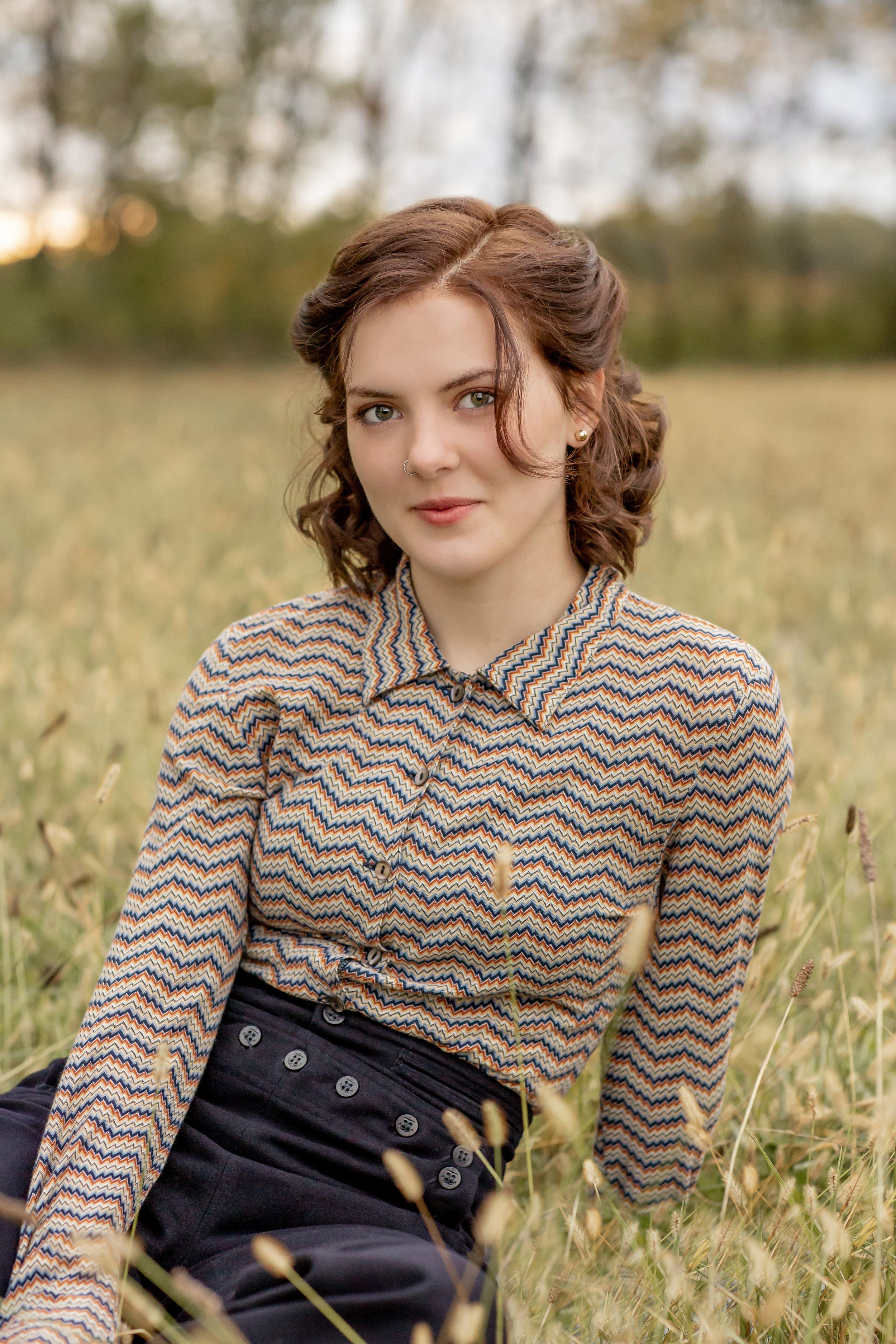 Young woman in patterned chevron shirt sitting in golden grass field during sunset portrait session.