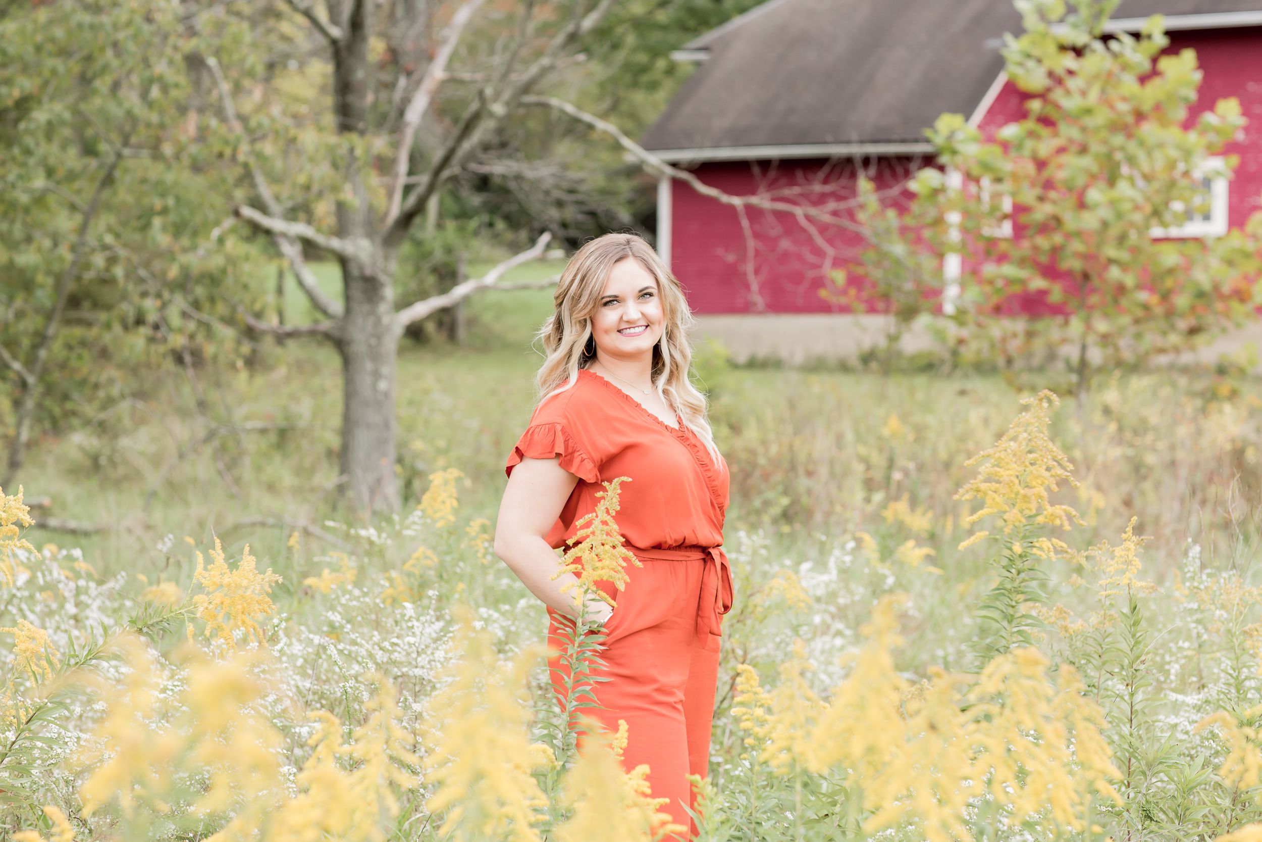 Woman in orange jumpsuit holding yellow flowers in a field with a red barn backdrop.