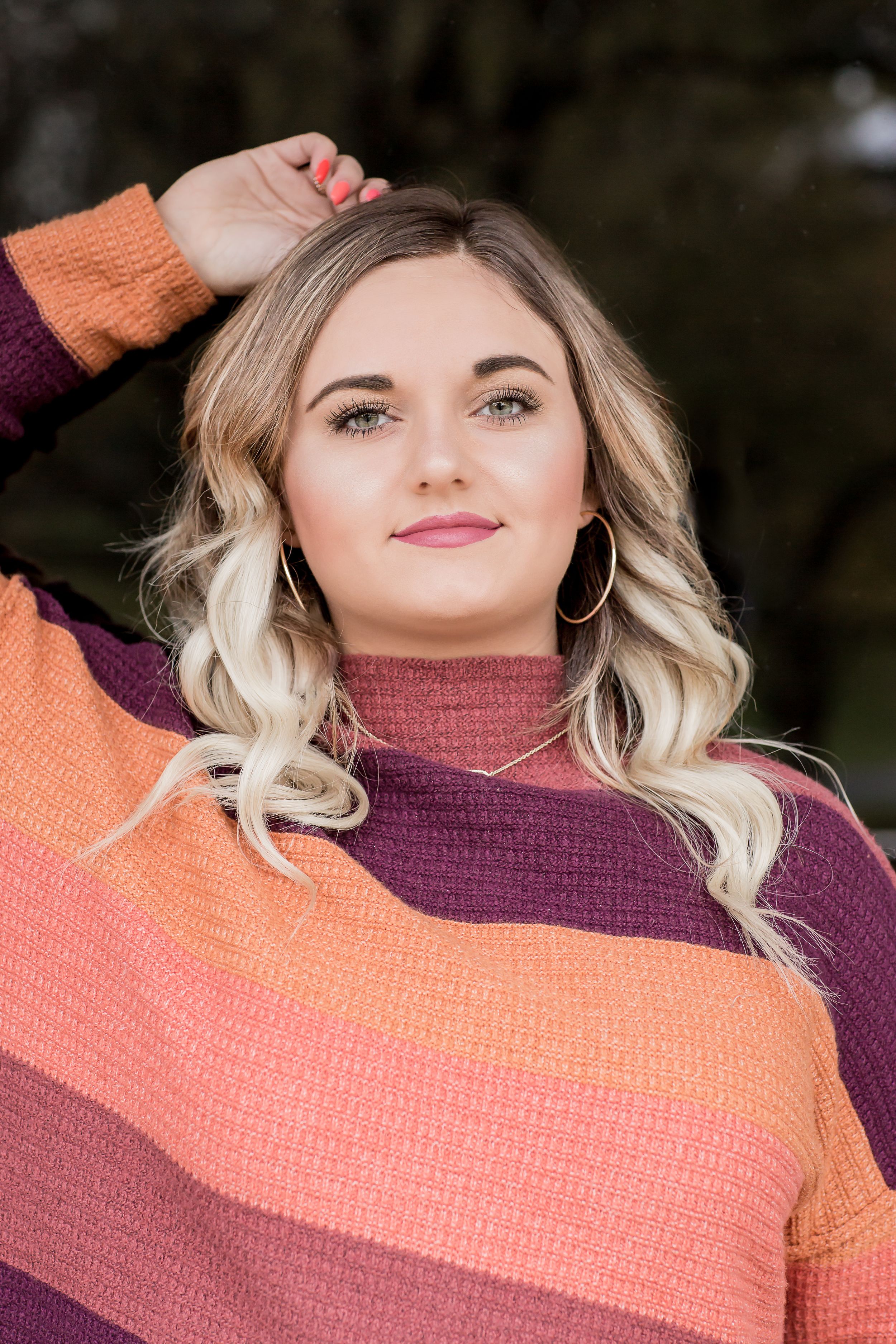 Young woman in colorful striped sweater poses outdoors with hand in blonde hair for senior portrait.