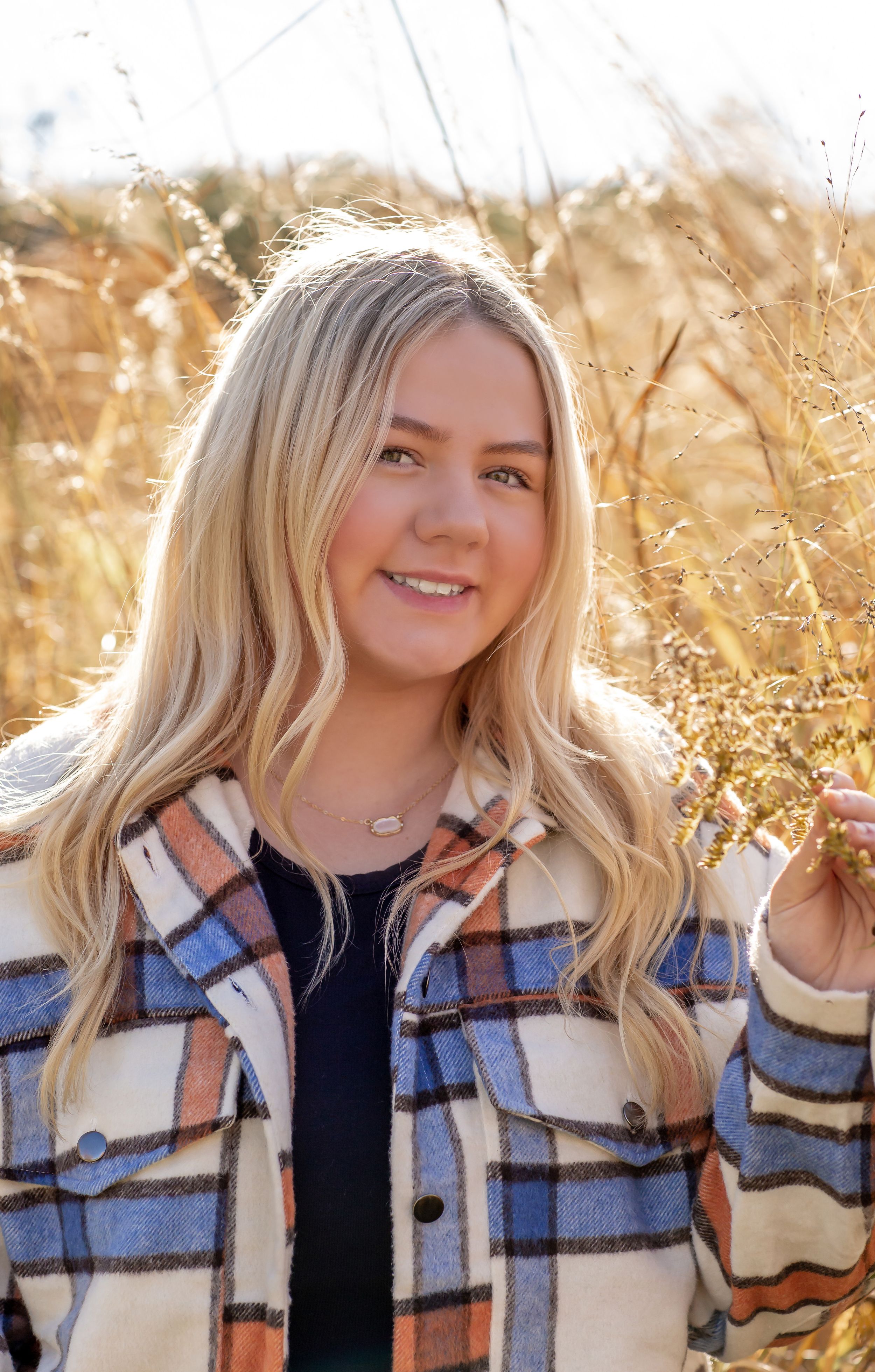 Smiling blonde young woman in plaid jacket standing in a golden field on a sunny day.