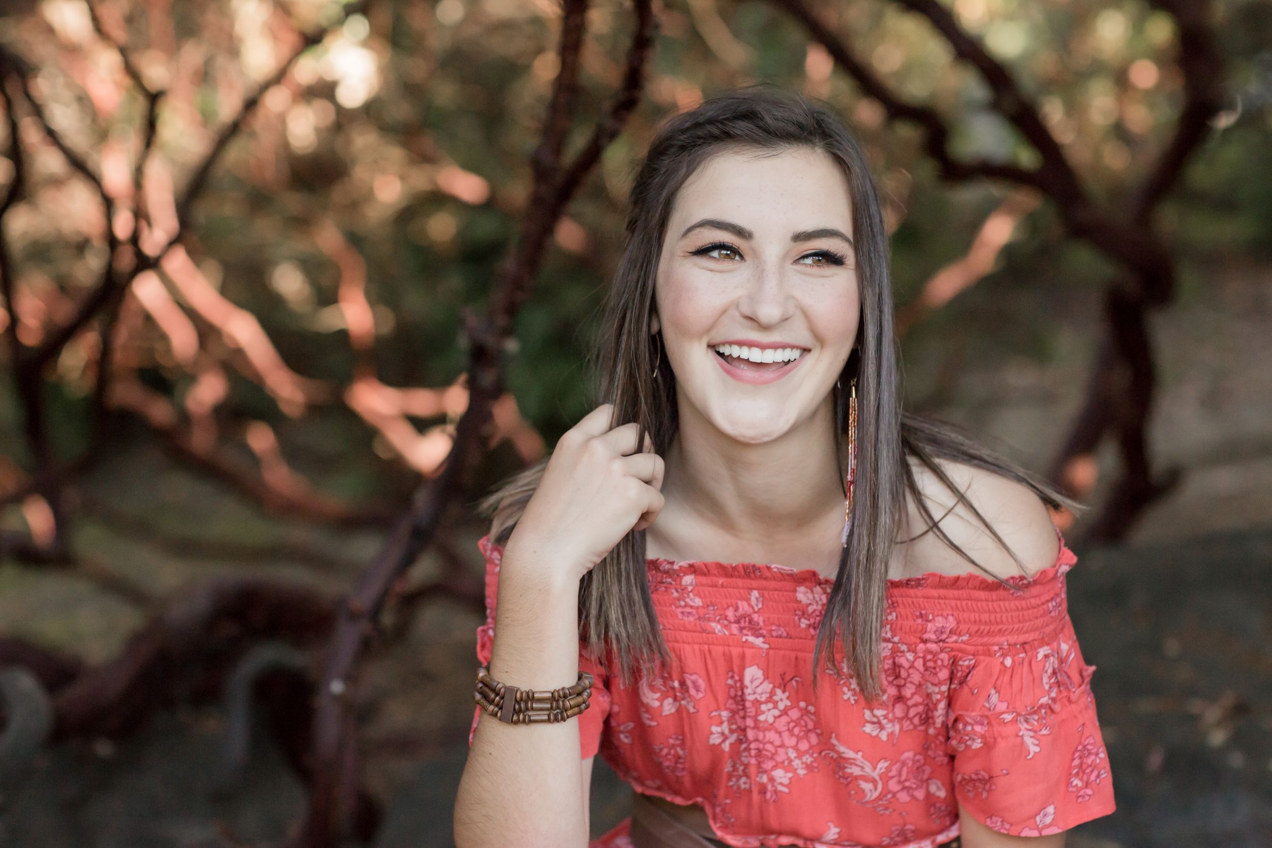 Smiling young woman in coral off-shoulder top laughs outdoors among red branches for senior photo.