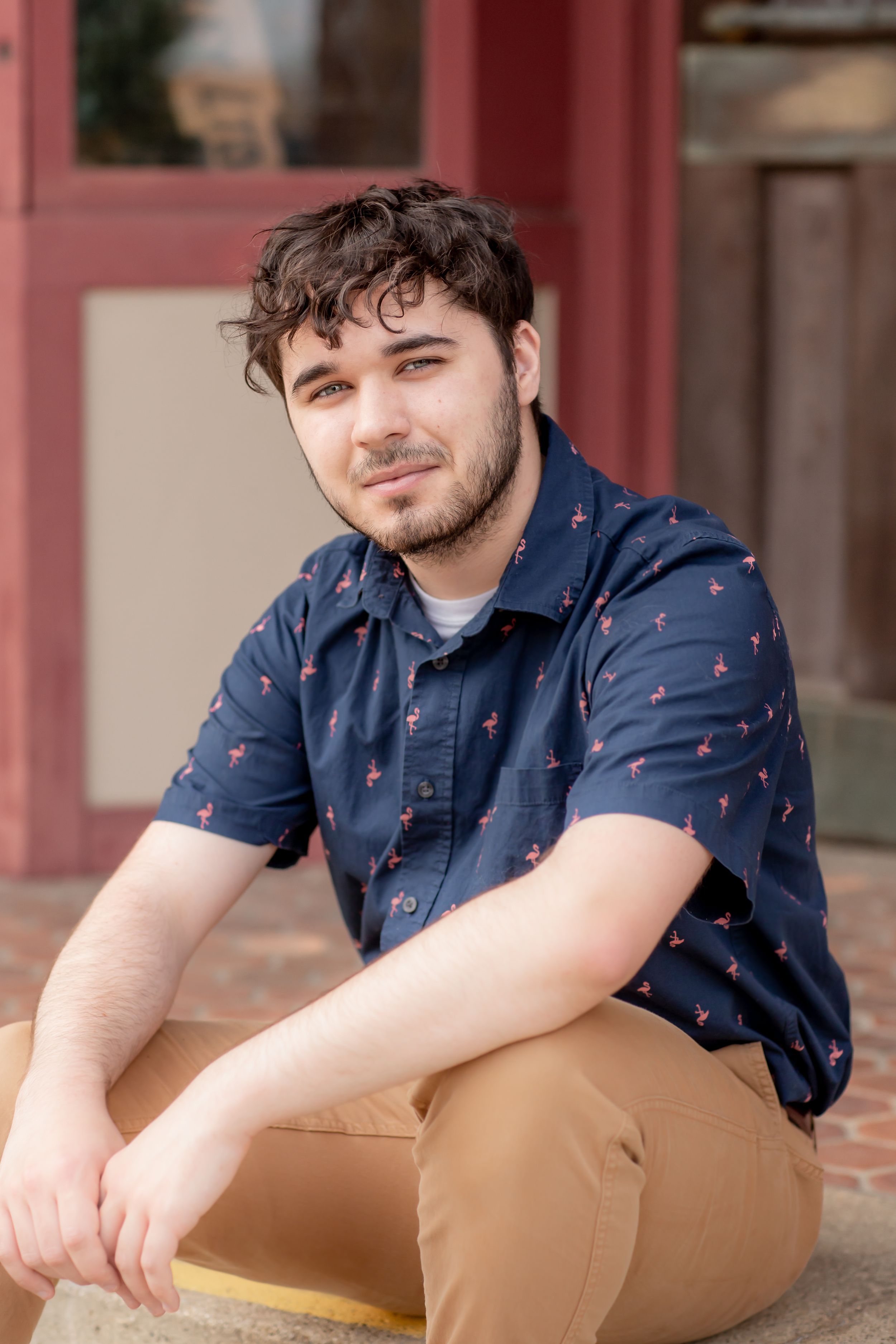 Young man with dark curly hair in navy flamingo print shirt sitting on outdoor steps.