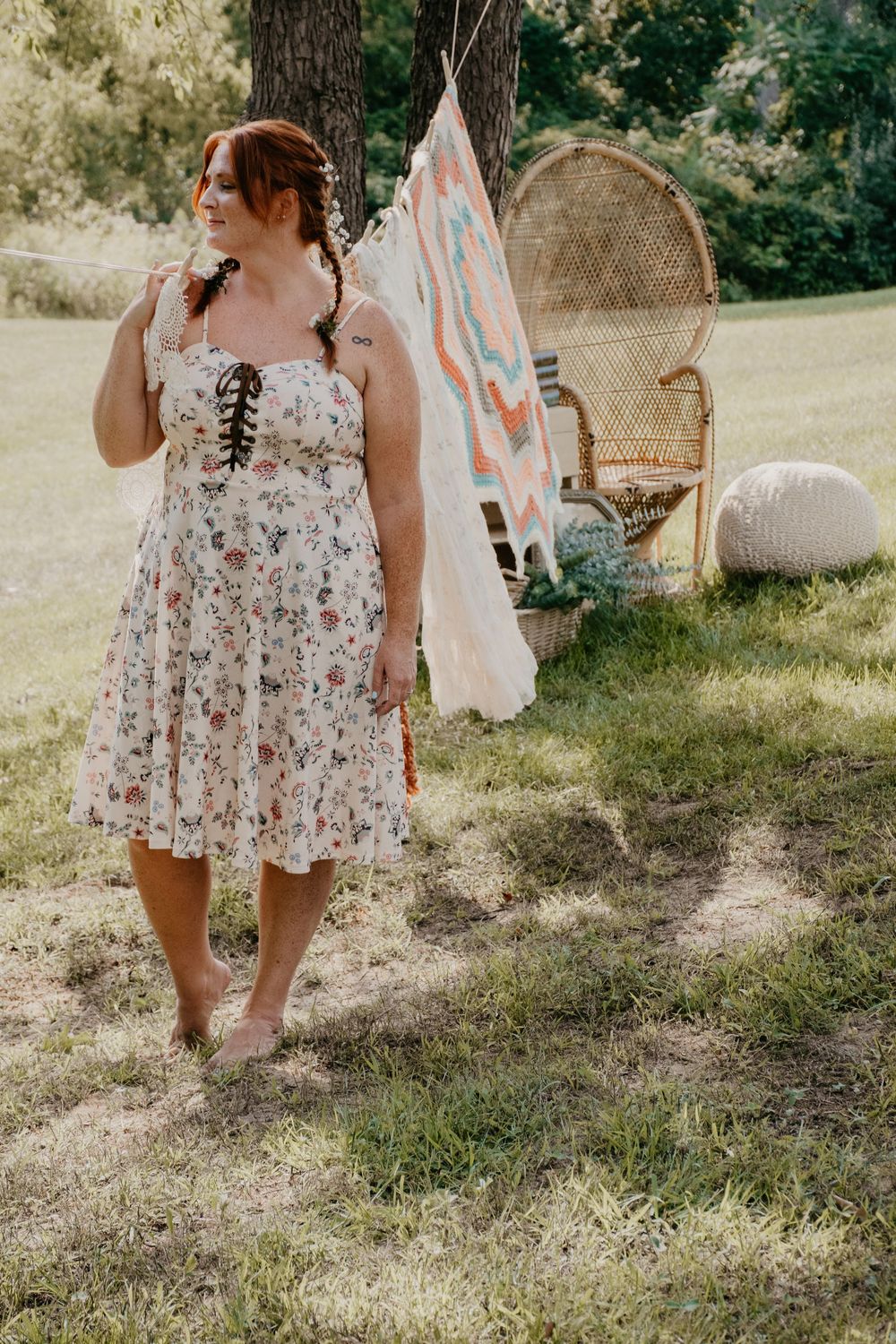 Redhead woman in floral sundress walking barefoot outdoors near boho teepee setup in green field.