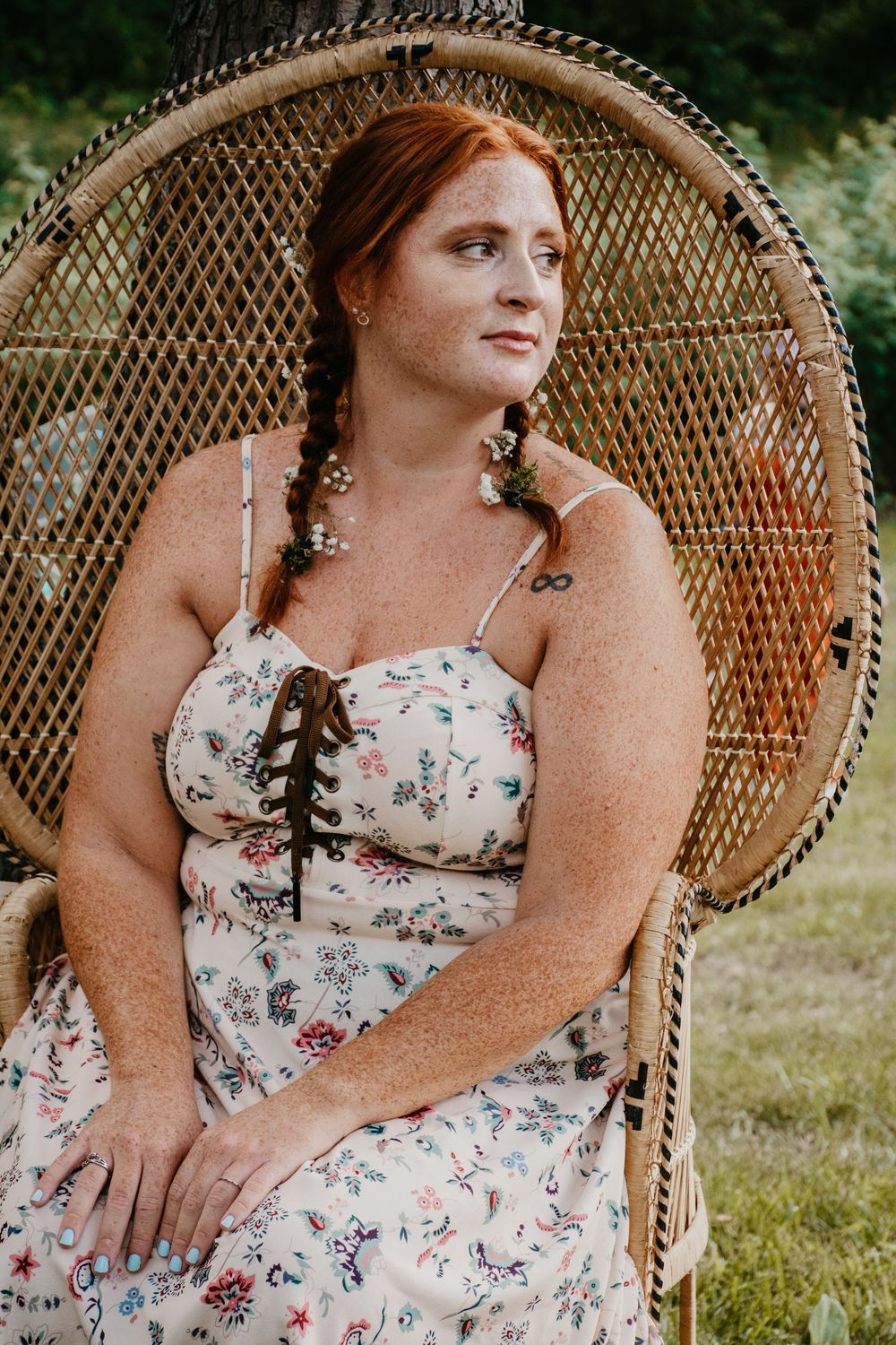 Freckled redhead woman in floral dress sitting in wicker peacock chair outdoors in natural light.