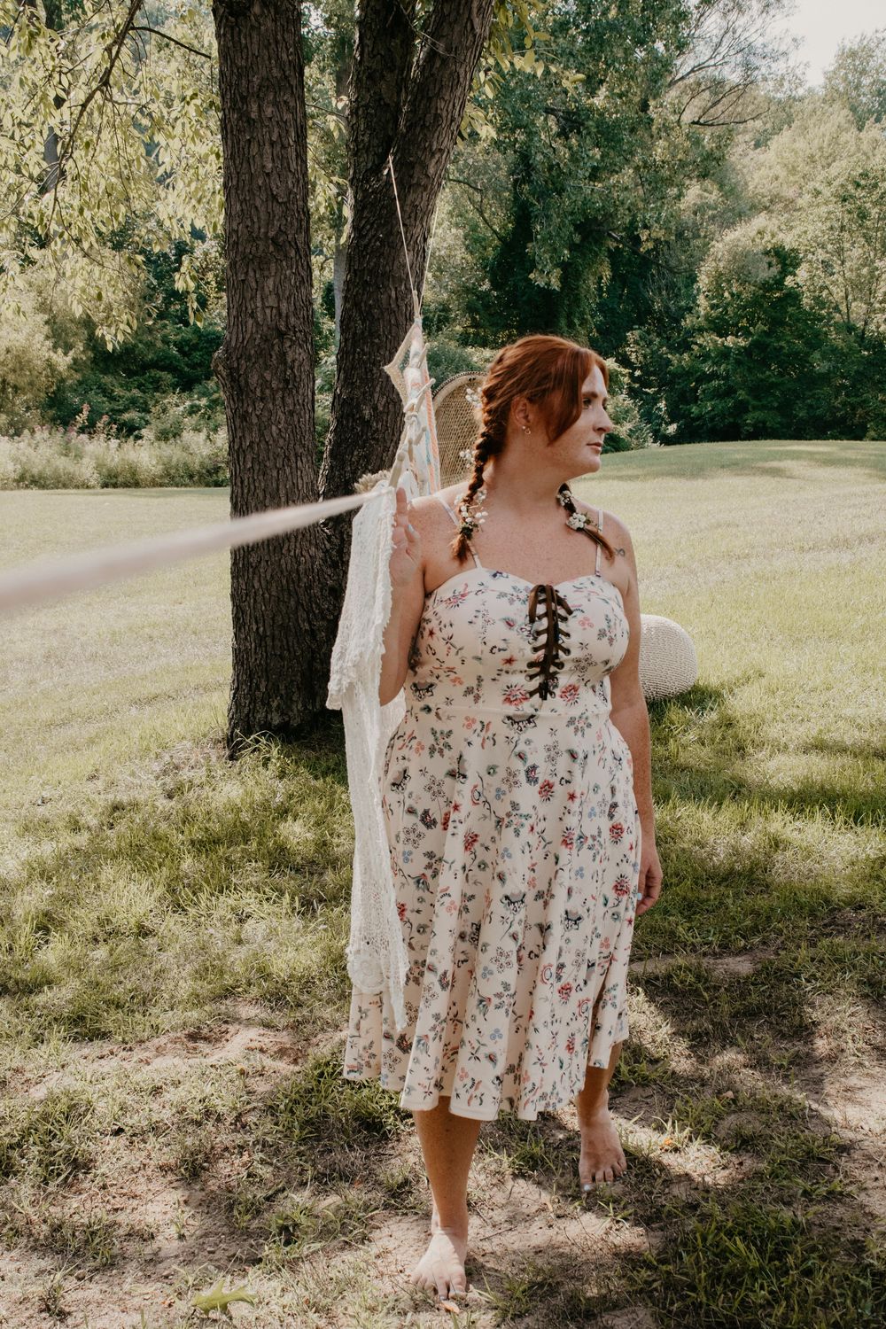 Barefoot redhead woman in floral dress walking under trees holding white lace garment outdoors.