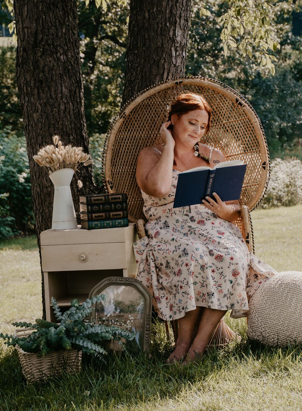 Woman in floral dress reading book in wicker peacock chair surrounded by vintage decor outdoors.