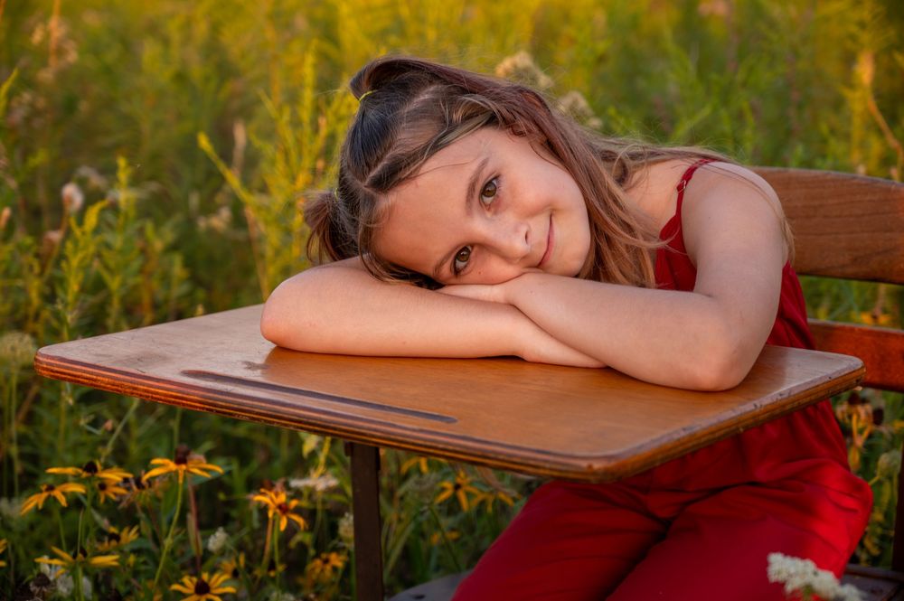 Young girl in red dress rests chin on hands at a wooden desk surrounded by a wildflower meadow.