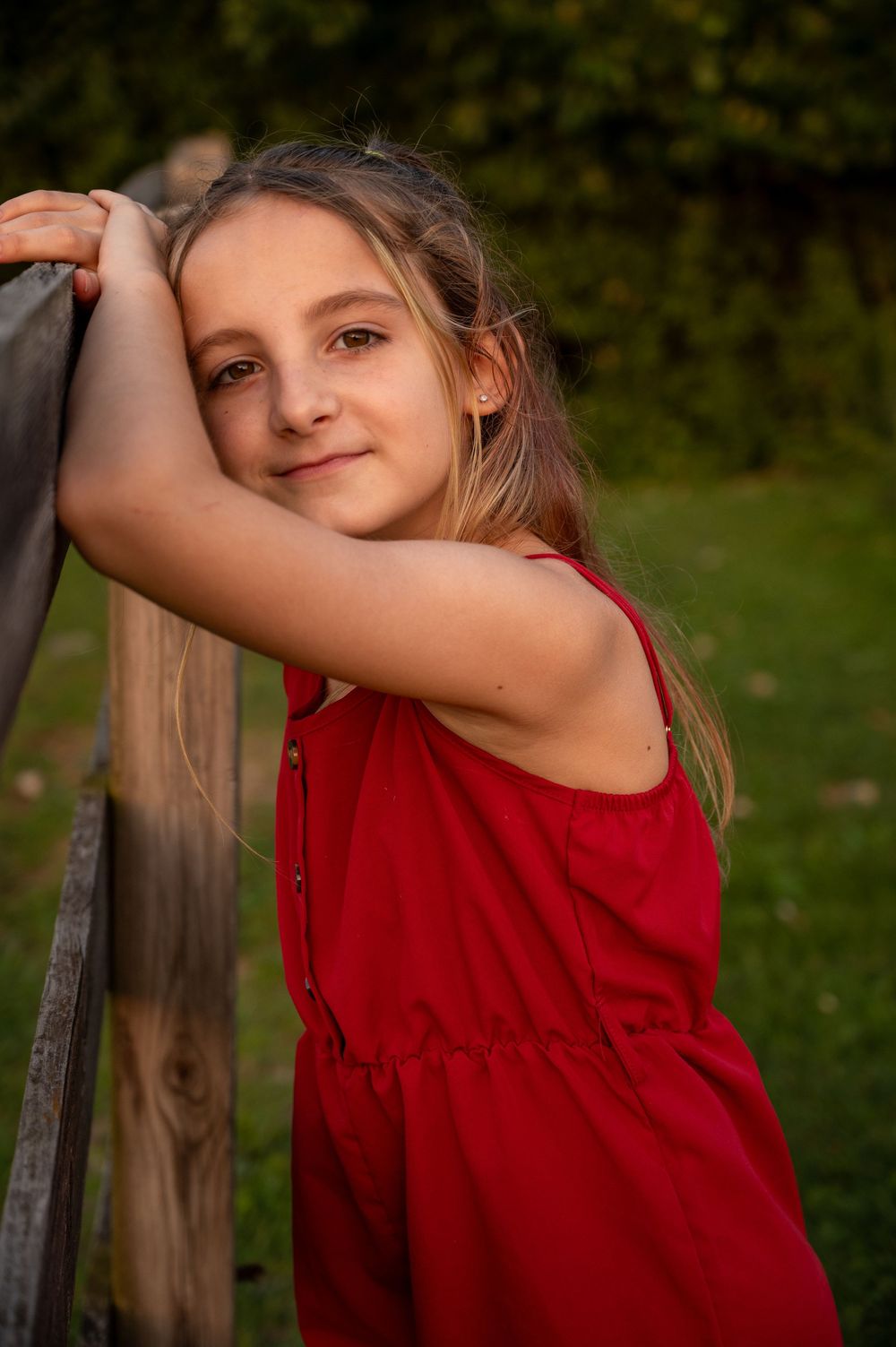 Young girl in red dress leaning on wooden fence outdoors, smiling softly in warm natural light.