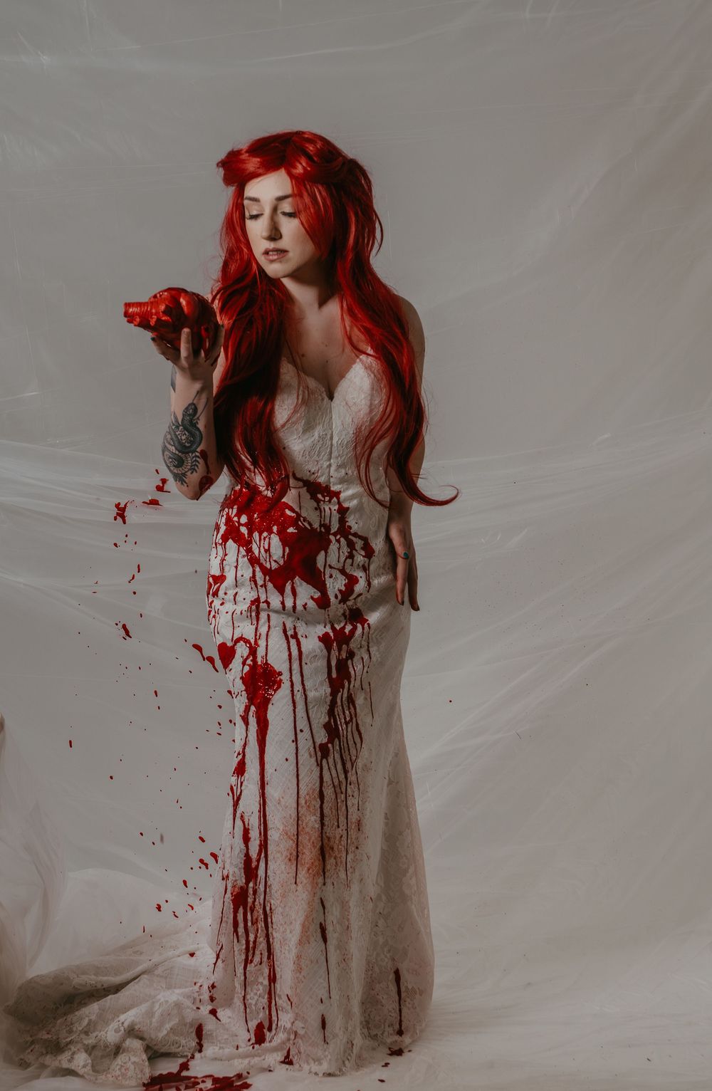 Woman with long red hair in blood-covered white wedding dress holds red object in studio shoot.