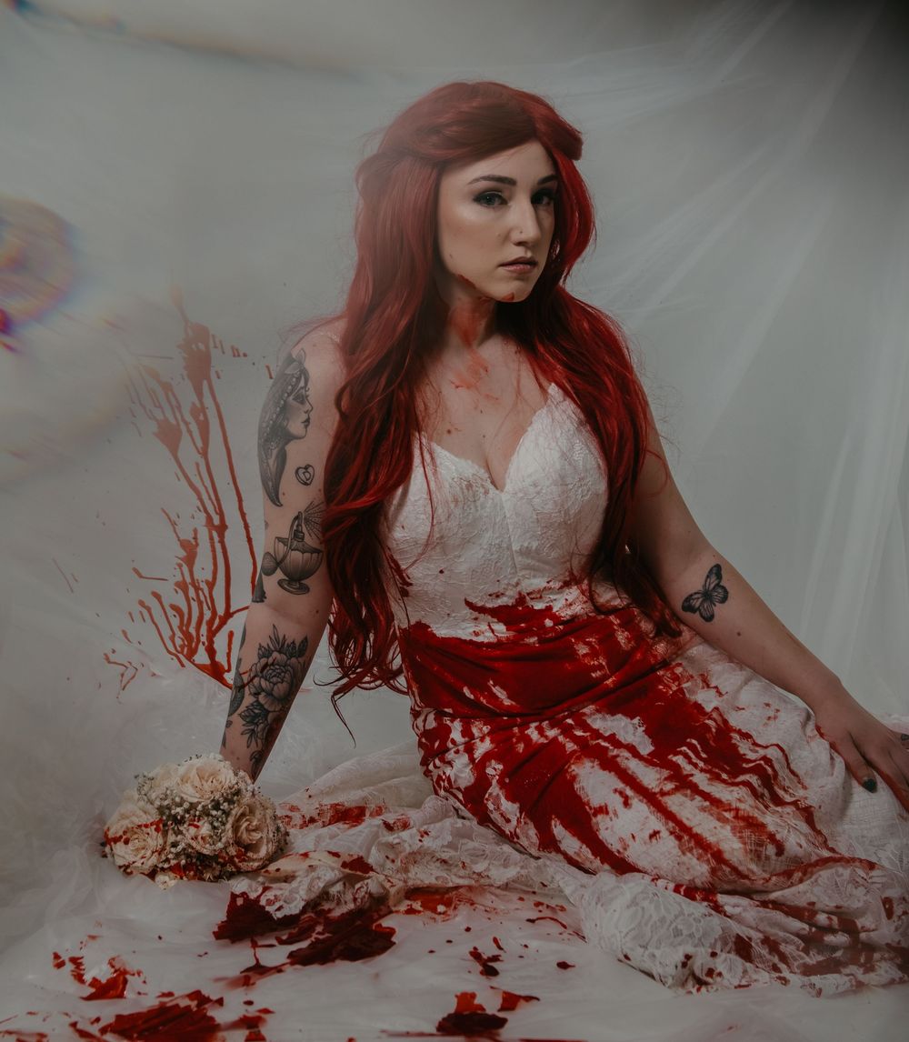 Woman in blood-splattered white wedding dress seated on floor in dramatic horror-themed photo shoot.