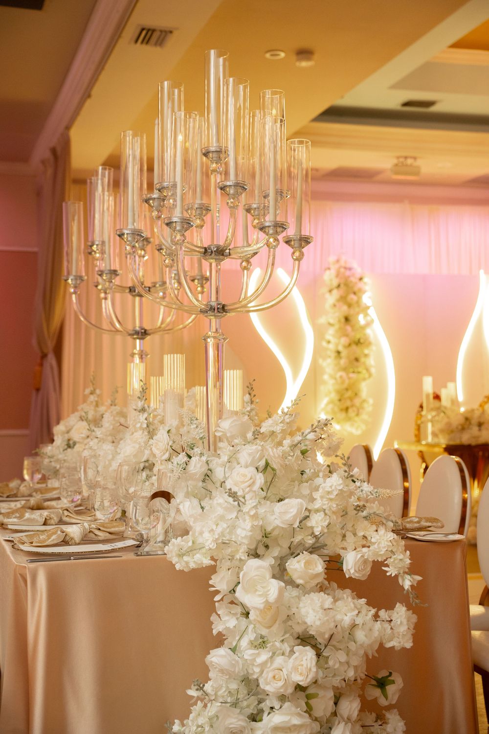 Elegant wedding reception table with white floral arrangements, tall glass candelabra, and blush pink decor in a luxurious ballroom.