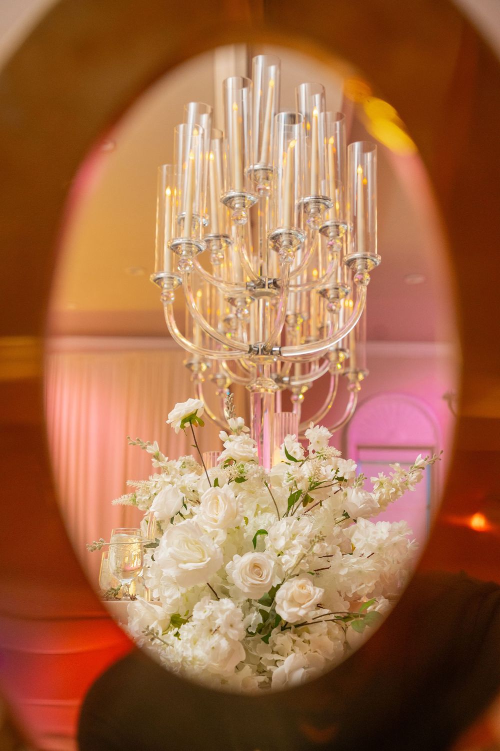 Elegant wedding centerpiece with crystal candelabra and white roses displayed through oval mirror frame.