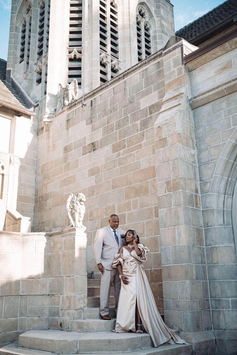 Elegant couple in formal attire posing on stone church steps, bride in satin gown, groom in light suit.