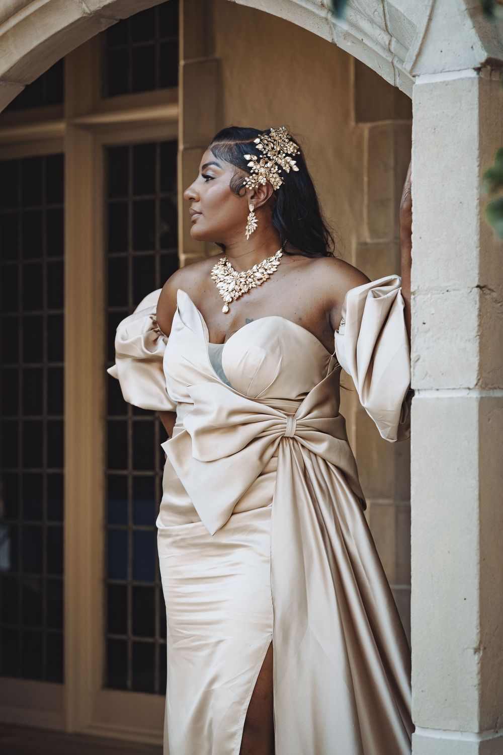 Elegant woman in champagne satin gown with bow detail, gold jewelry and hair piece, posing by stone archway.