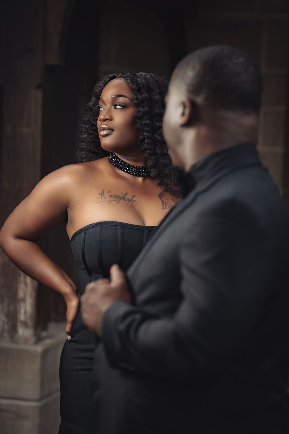 A Black couple in elegant black attire pose intimately in a dimly lit setting, the woman wearing a strapless gown and choker.