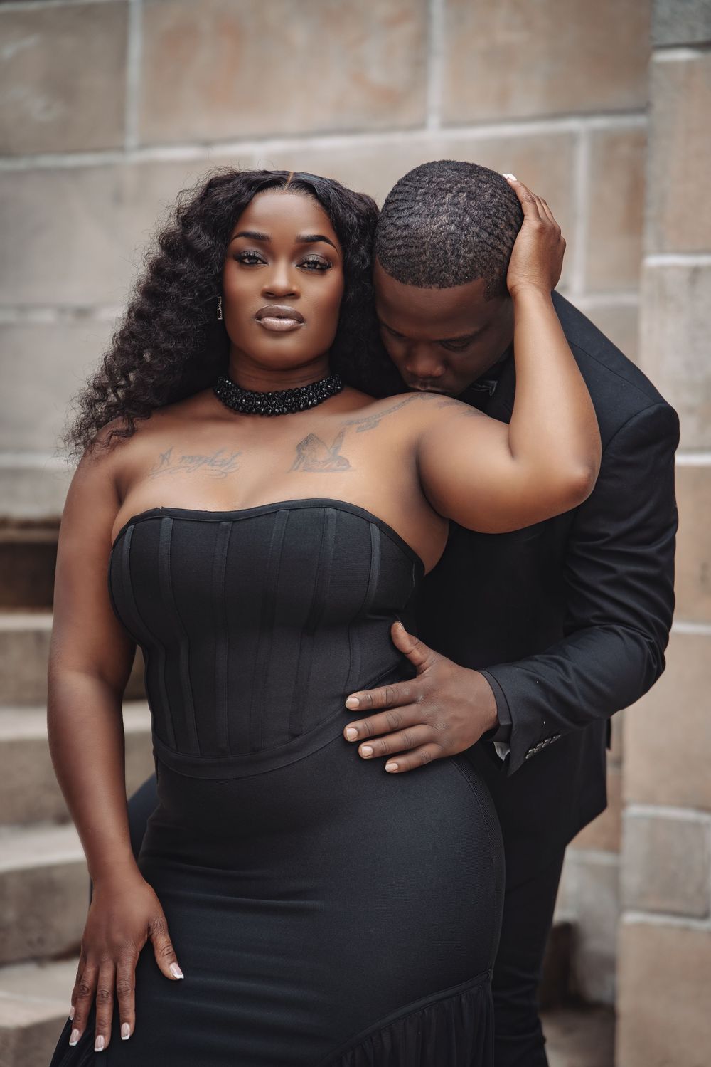 A couple in elegant black outfits poses intimately against a stone wall, the man nuzzling the woman's shoulder.