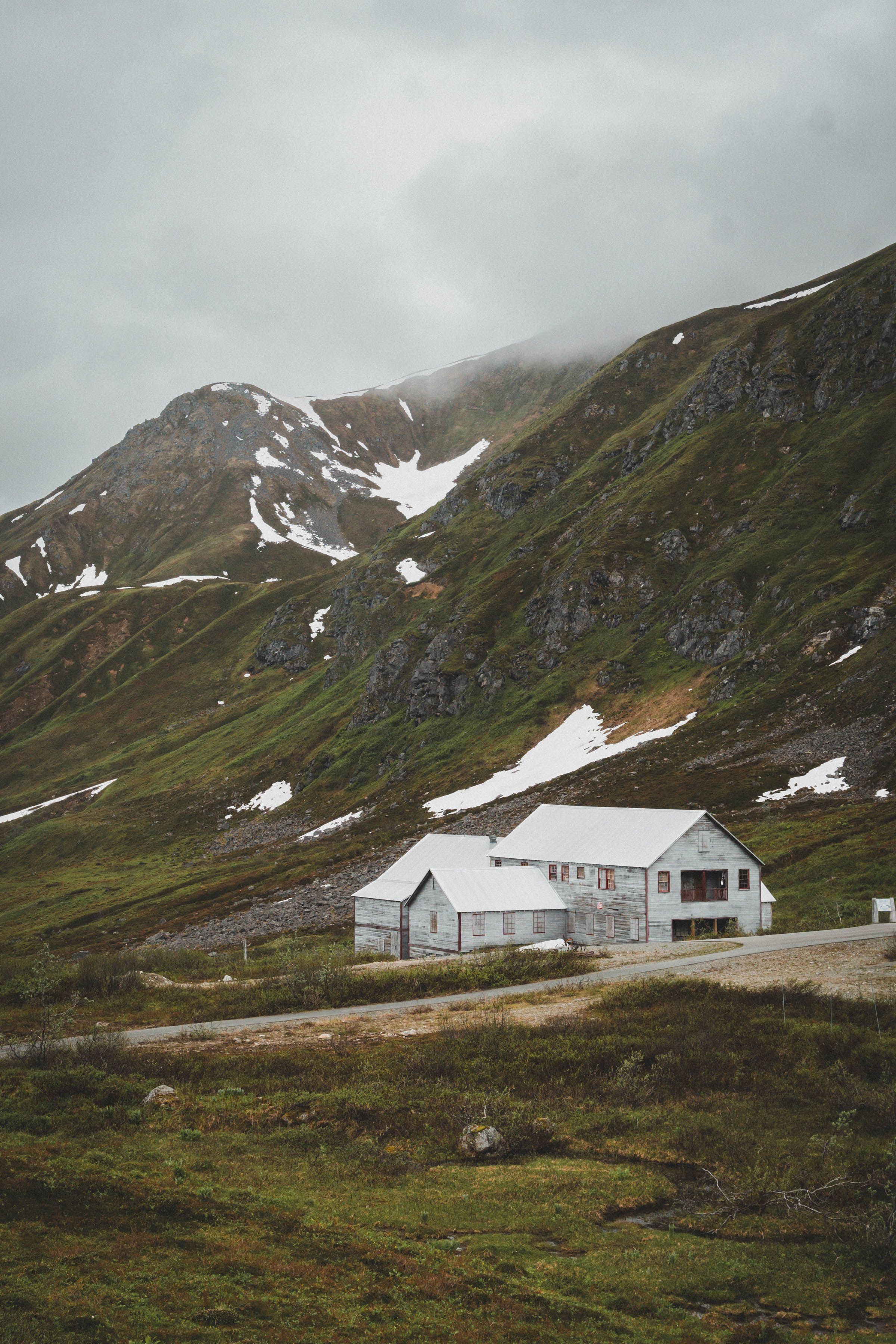 A remote white house sits at the base of a snow-patched green mountain under cloudy skies.