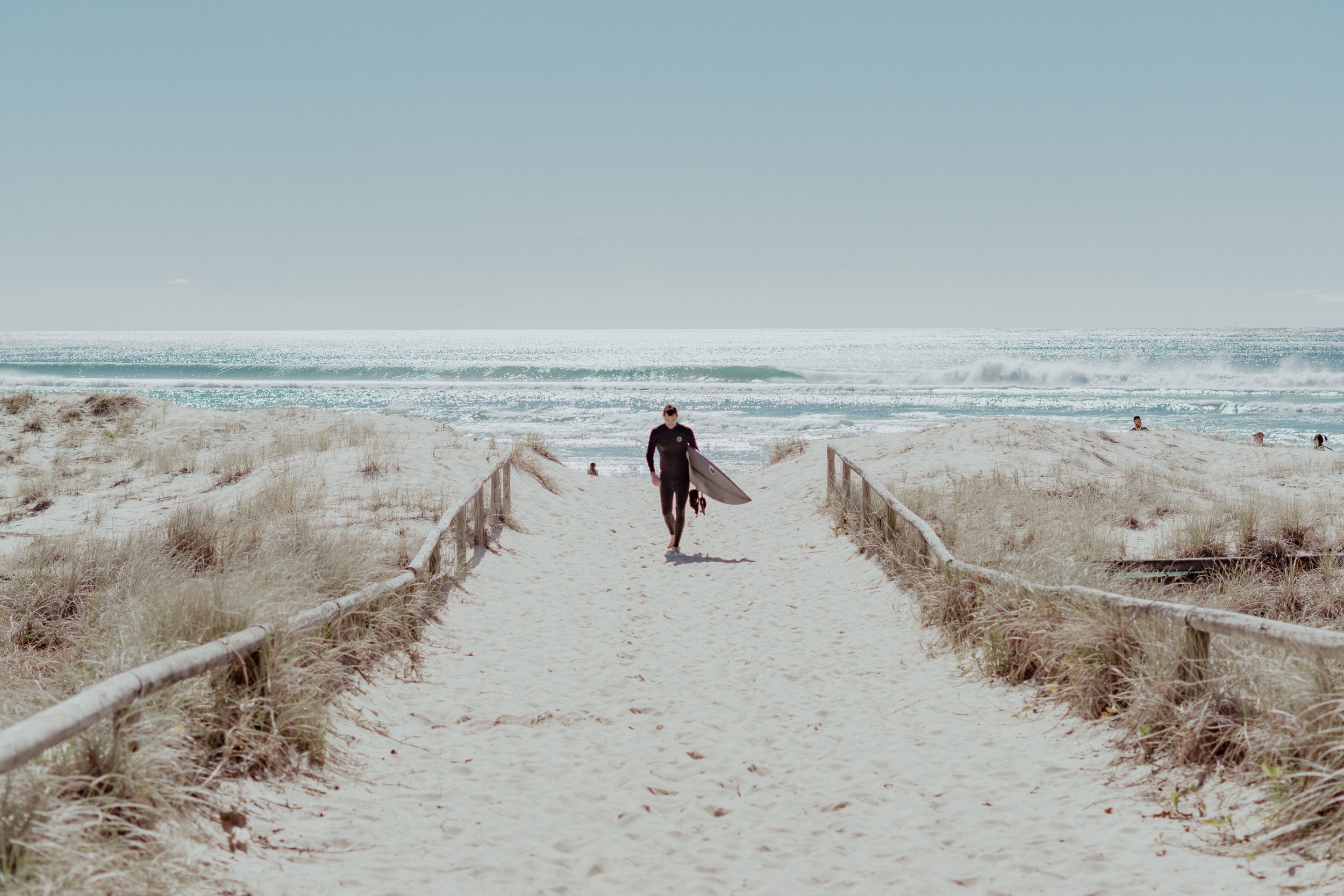 Surfer walking down a sandy path toward ocean waves on a clear day.