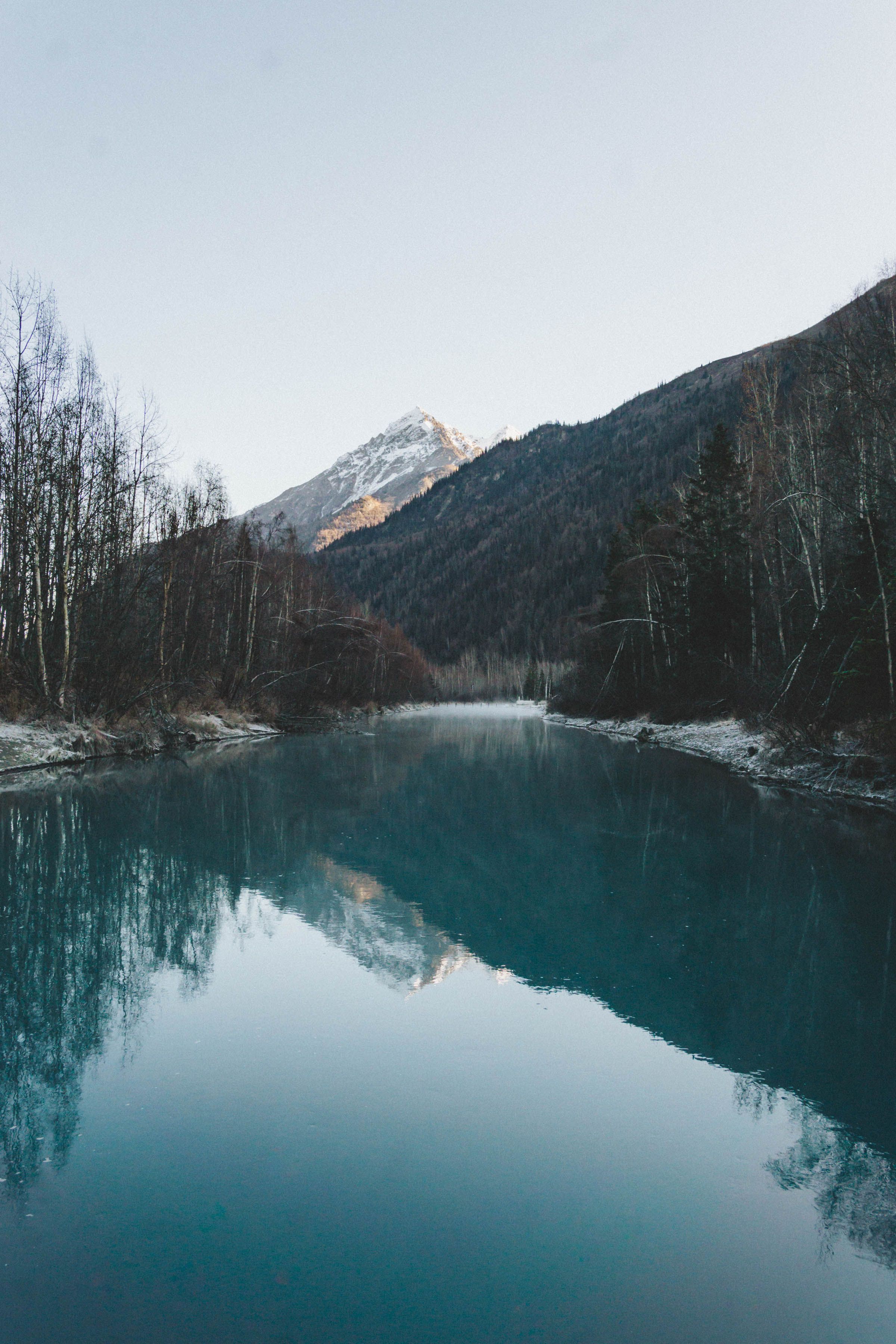 A serene turquoise lake reflects snow-capped mountains surrounded by bare winter trees at dusk.