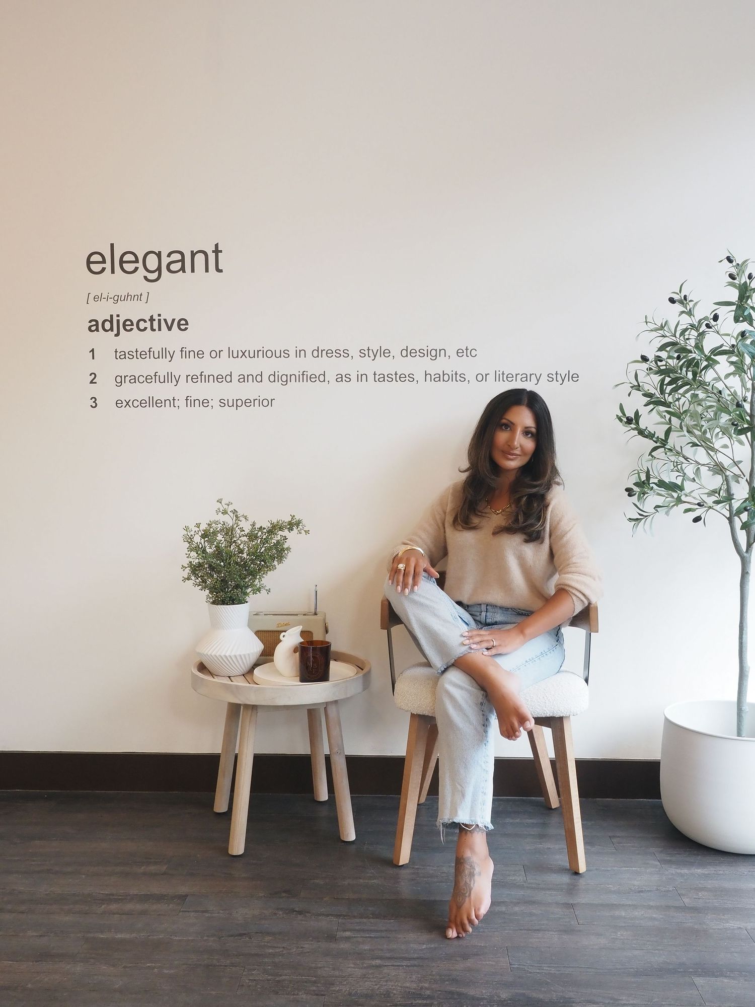 A casual-chic woman stands in a bright modern hallway with large indoor plants and glass doors.