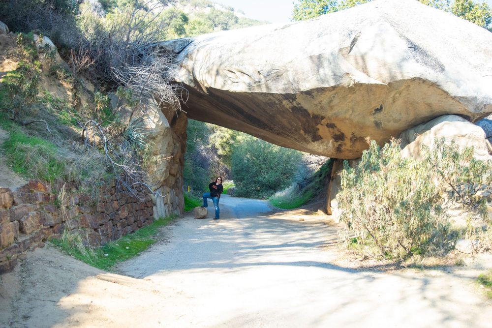 Large natural rock formation creates archway over hiking trail in mountainous landscape.