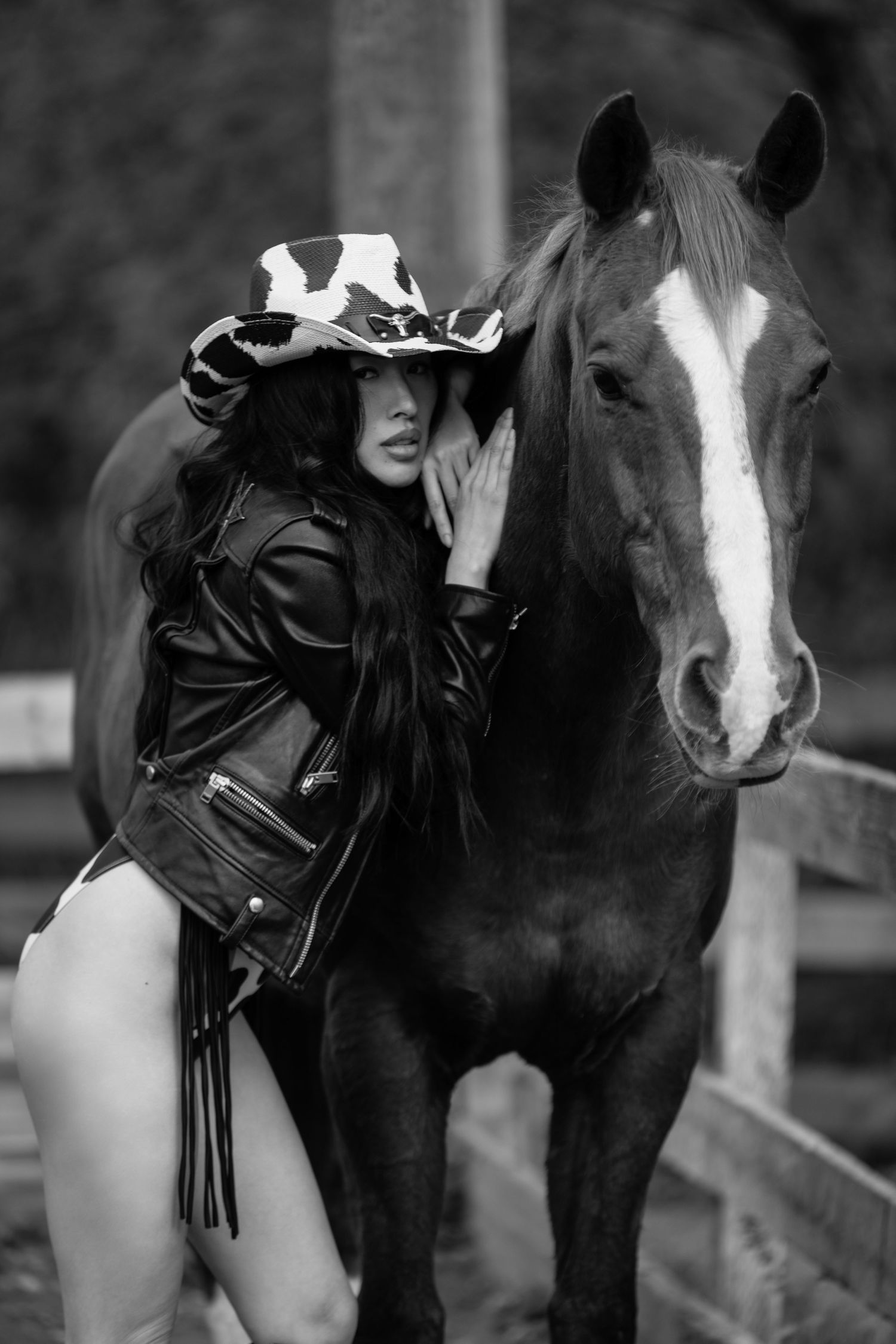 Black and white photo of a person in a leather jacket and hat standing next to a horse with a white blaze marking.