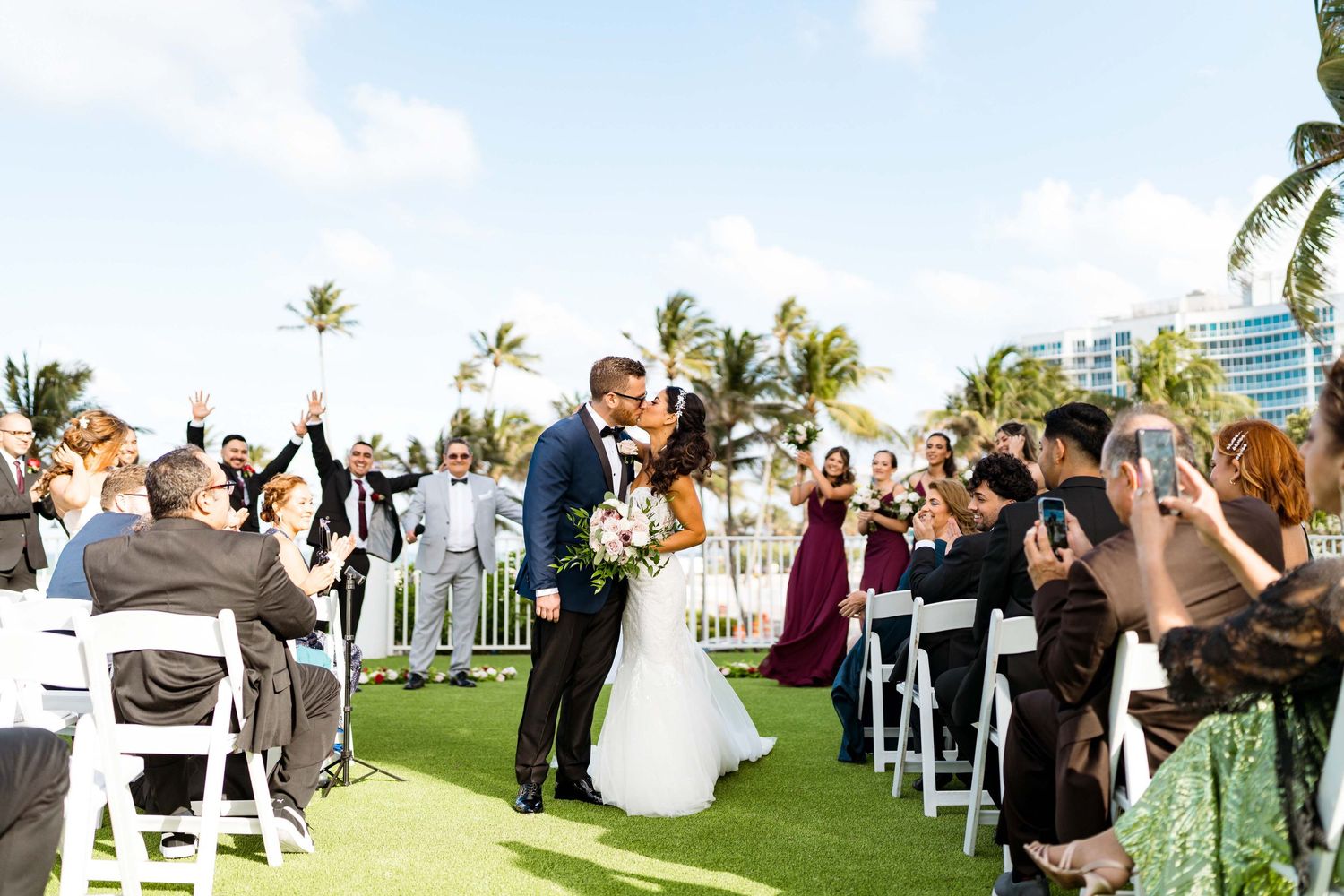 Outdoor wedding ceremony on a tropical lawn with palm trees and white chairs.