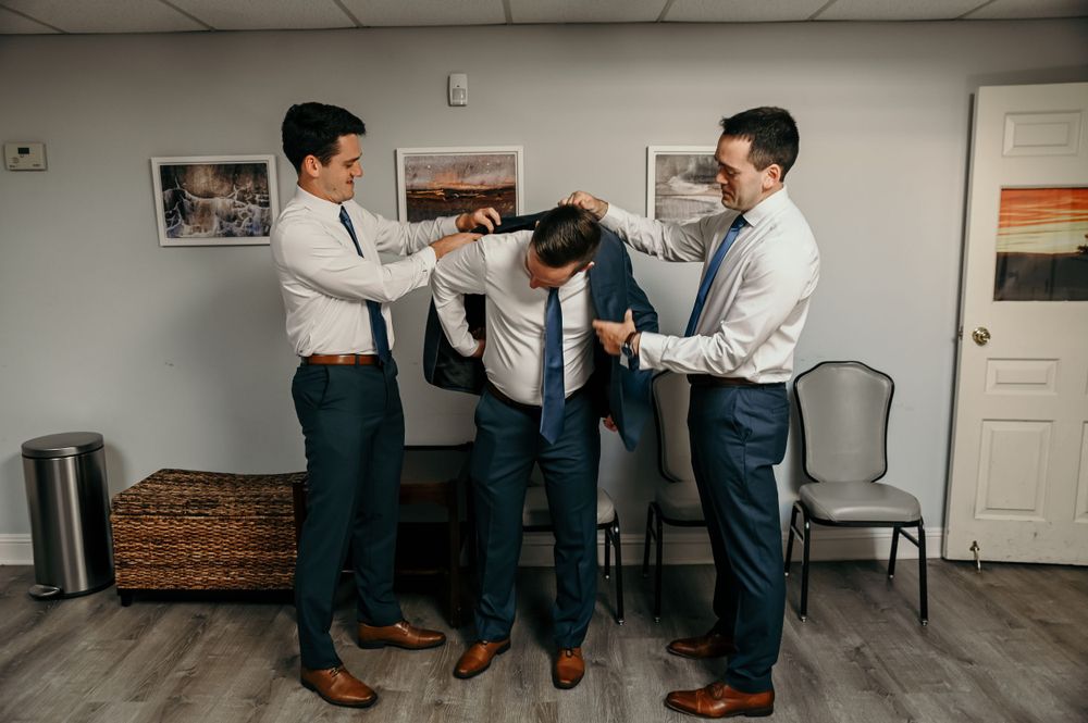 A group in navy suits adjusts ties and gets ready in a bright indoor space.