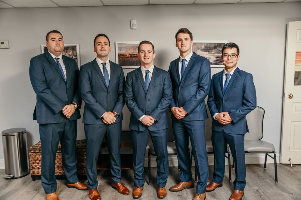 Wedding party members in blue suits pose together against a white wall.
