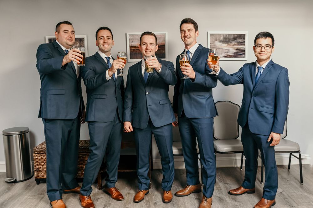 Wedding party members in navy suits pose together with drinks in a casual setting.