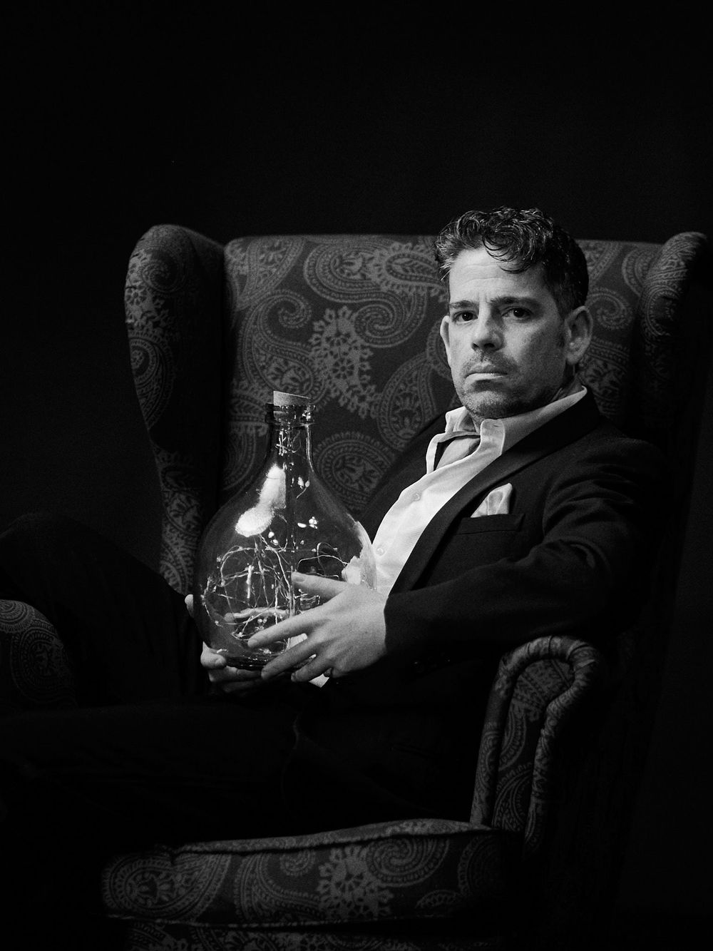 Eccentric traditional portrait of a man dressed in a suit, sitting in a wingback chair, holding a glass jar