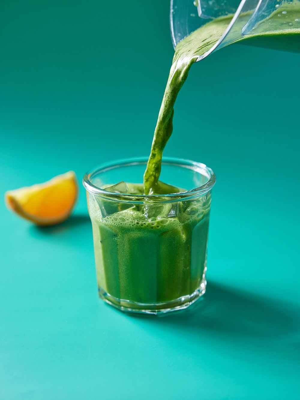 A green juice being poured into a glass on a green background with an orange slice to the side