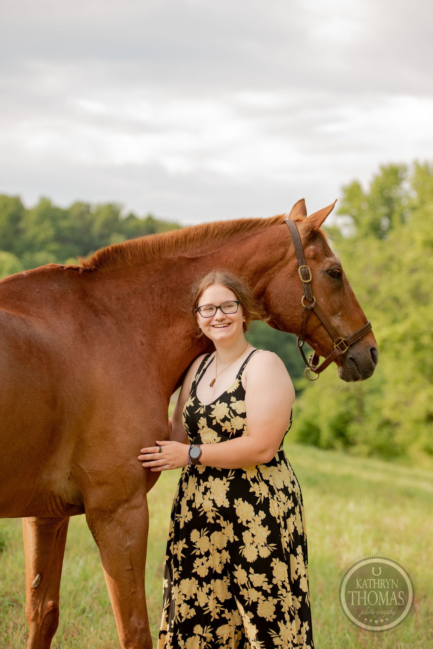 A beautiful chestnut horse poses with someone wearing a black and gold floral dress in a green field.